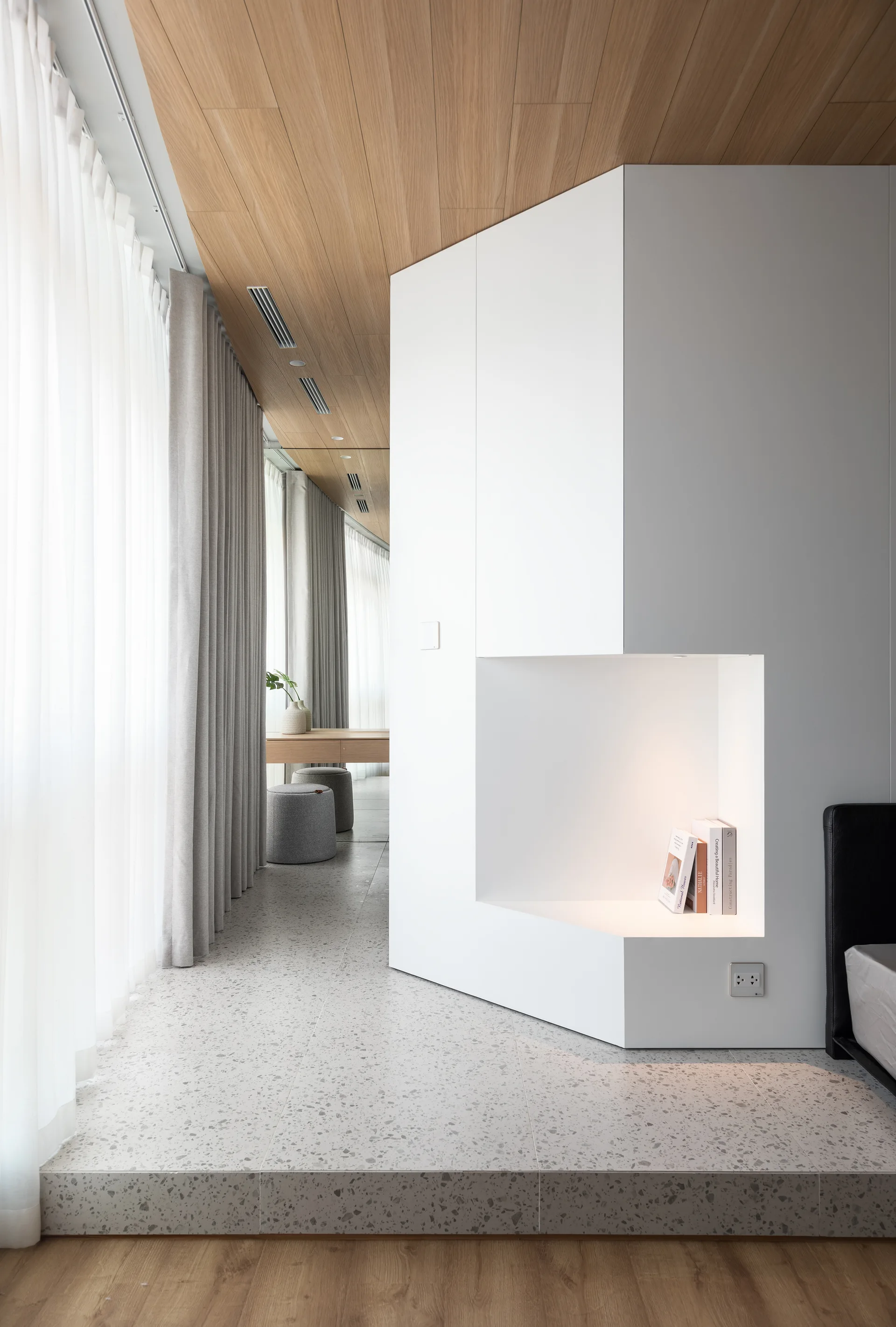 A minimalist bedroom features a custom white built-in unit with an illuminated niche, a wood-paneled ceiling, and speckled terrazzo flooring.