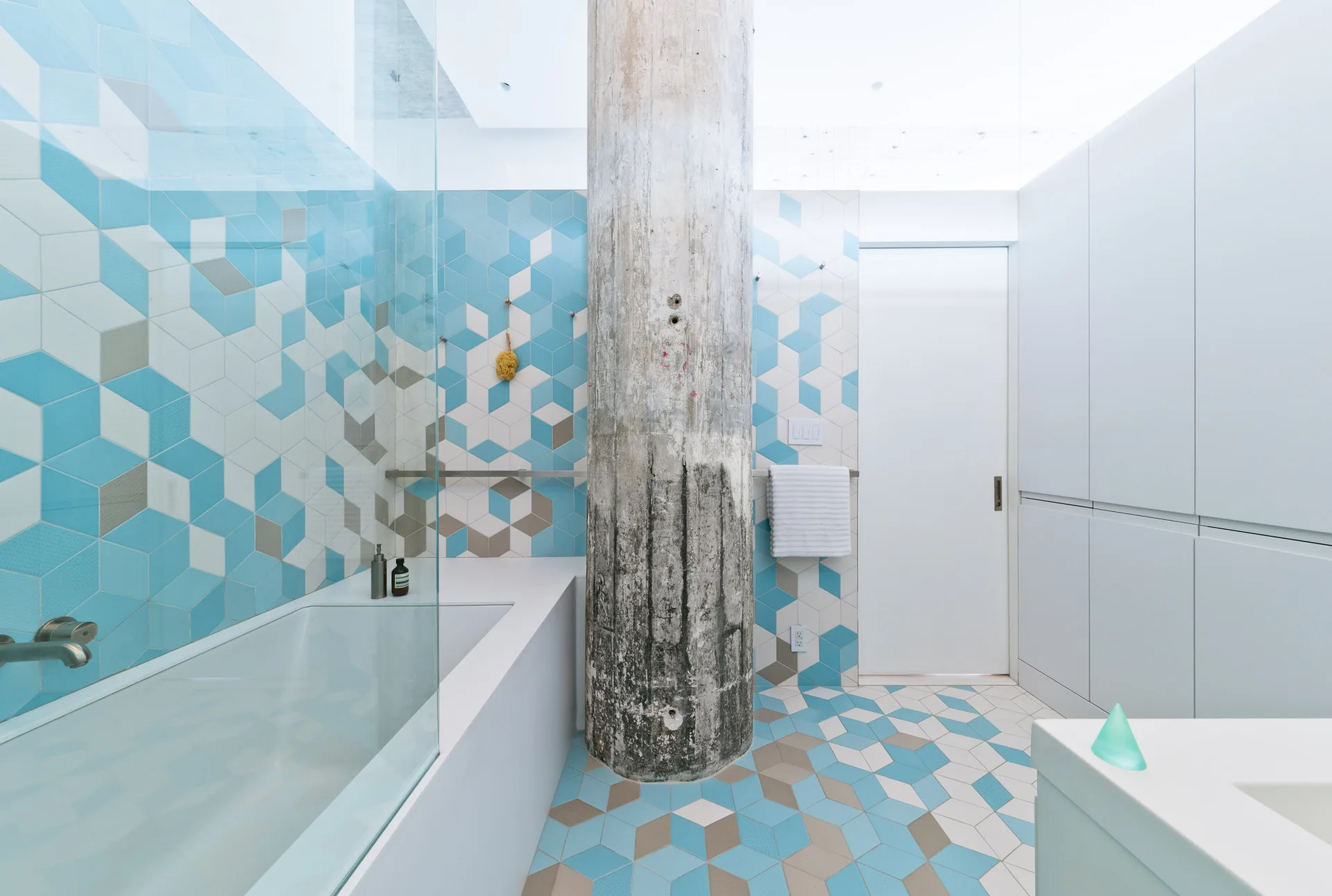 A modern bathroom features a prominent raw concrete column, a built-in bathtub with a glass partition, and blue, white, and grey geometric tiles.