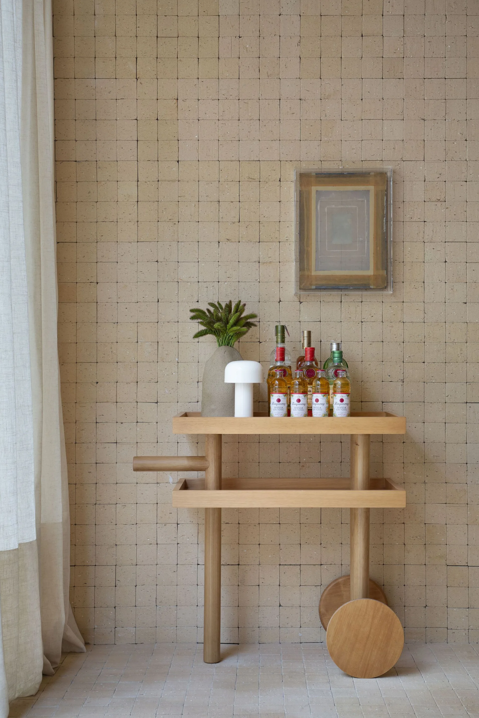 A light wood bar cart with round wheels holds spirits and a small white lamp against a wall of square beige tiles.