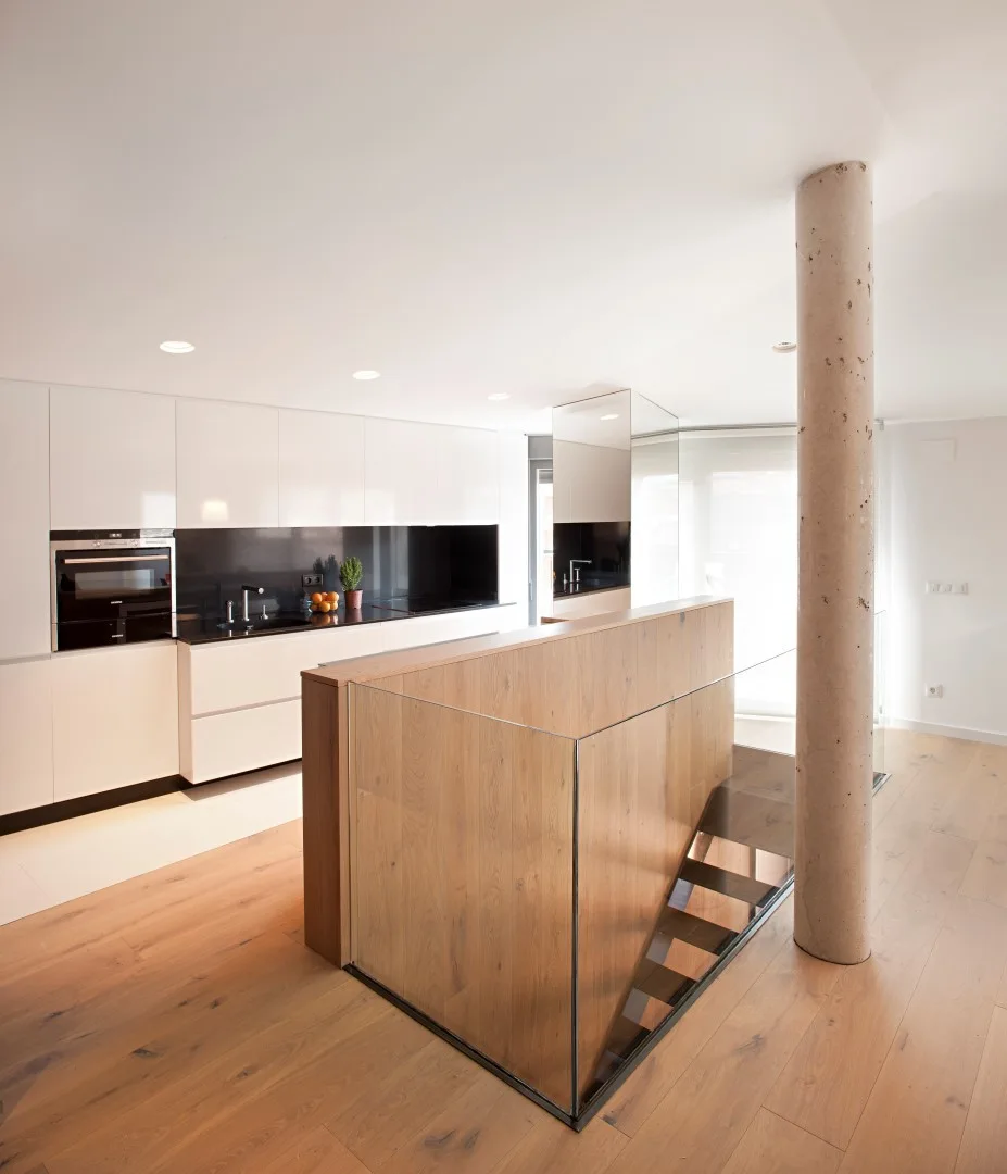A minimalist white kitchen with a black backsplash features a wood staircase with a glass railing and a concrete structural pillar on light wood floors.