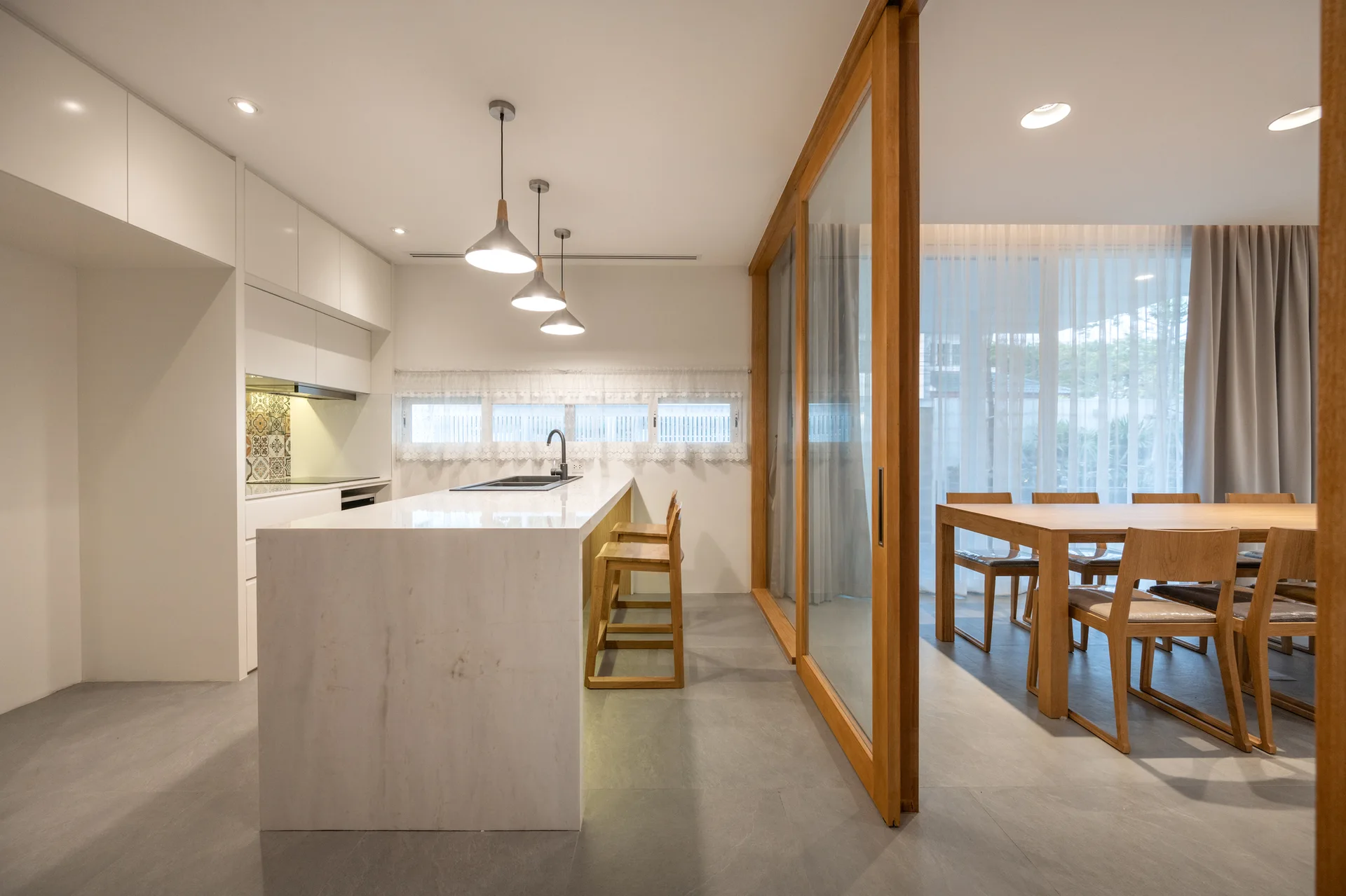 A modern white kitchen with a stone island and wood bar stools is separated from a dining area by glass sliding doors.