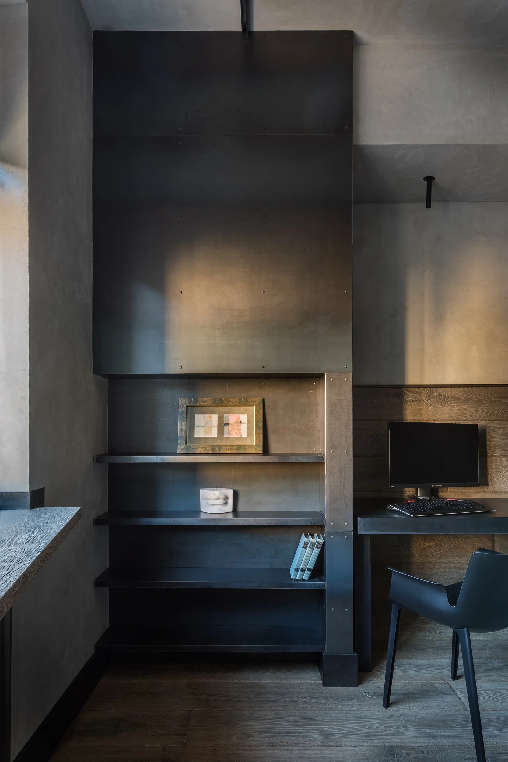 A modern industrial home office features a dark metal shelving unit, a black desk with a computer, wood paneled walls, and a black molded chair.
