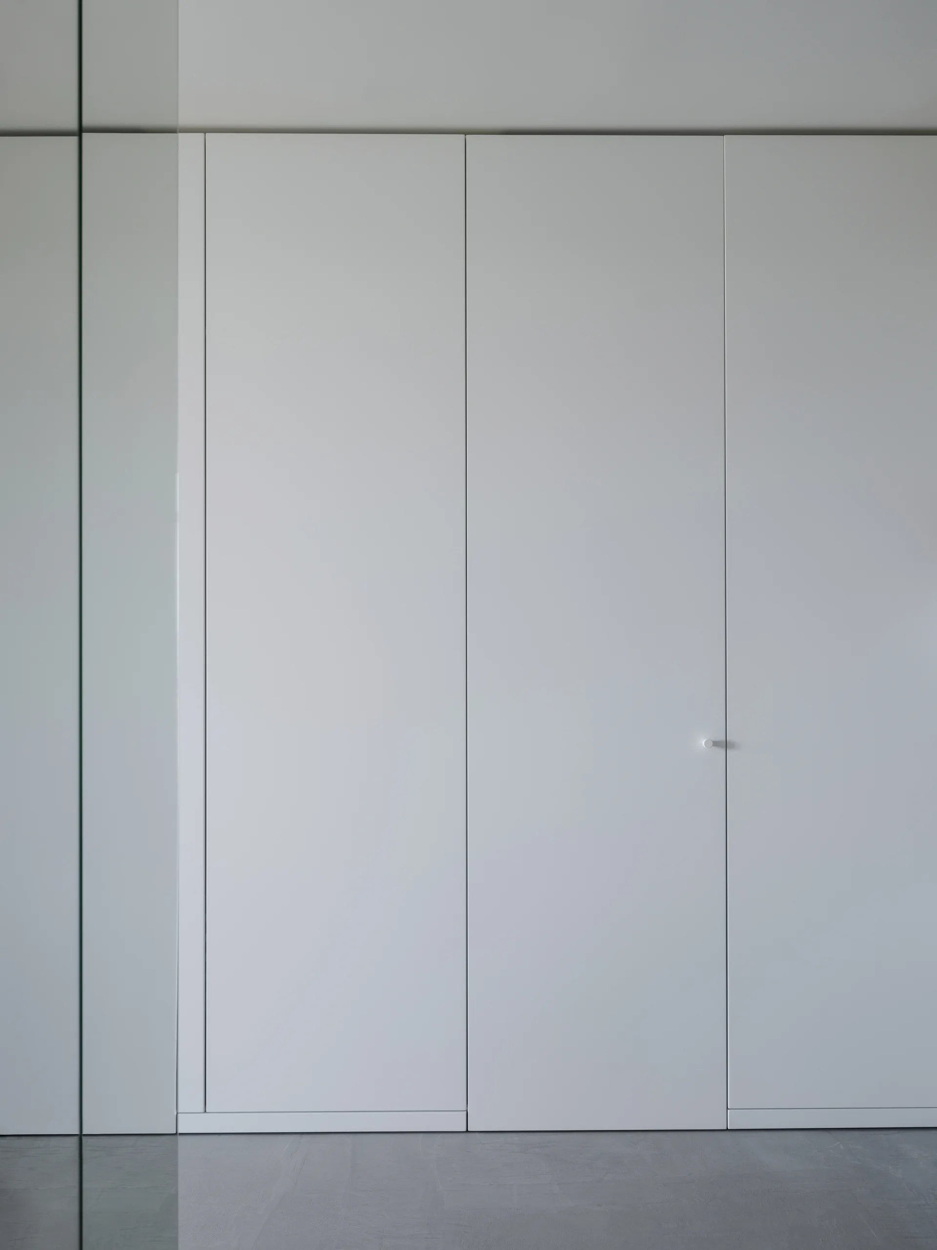 A floor-to-ceiling minimalist white closet features smooth matte panels, vertical seams, and a small cylindrical white door knob on a grey floor.