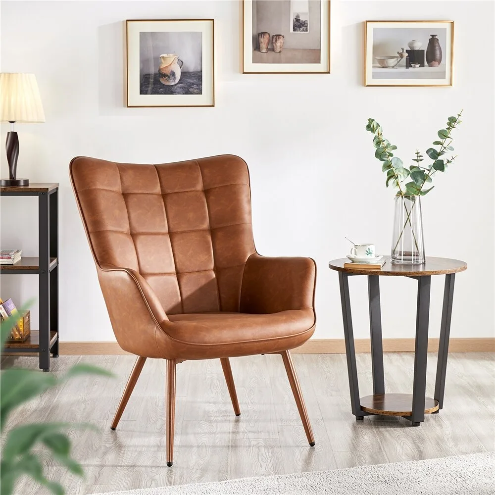 A tufted brown leather armchair sits next to a small round wooden side table in a room with light wood floors and white walls decorated with framed art.