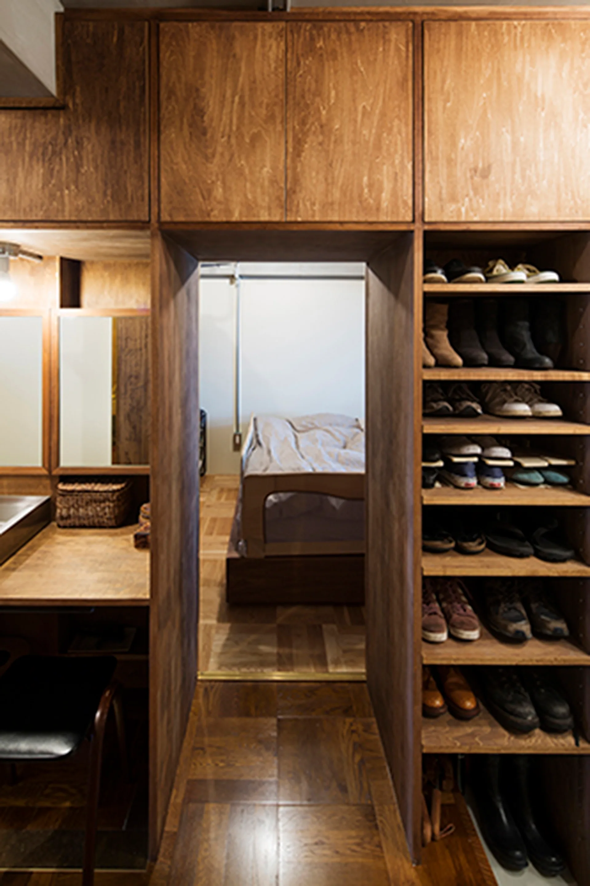 A wooden built-in storage unit with a central doorway connects a vanity area and a shoe rack to a bedroom with parquet flooring.