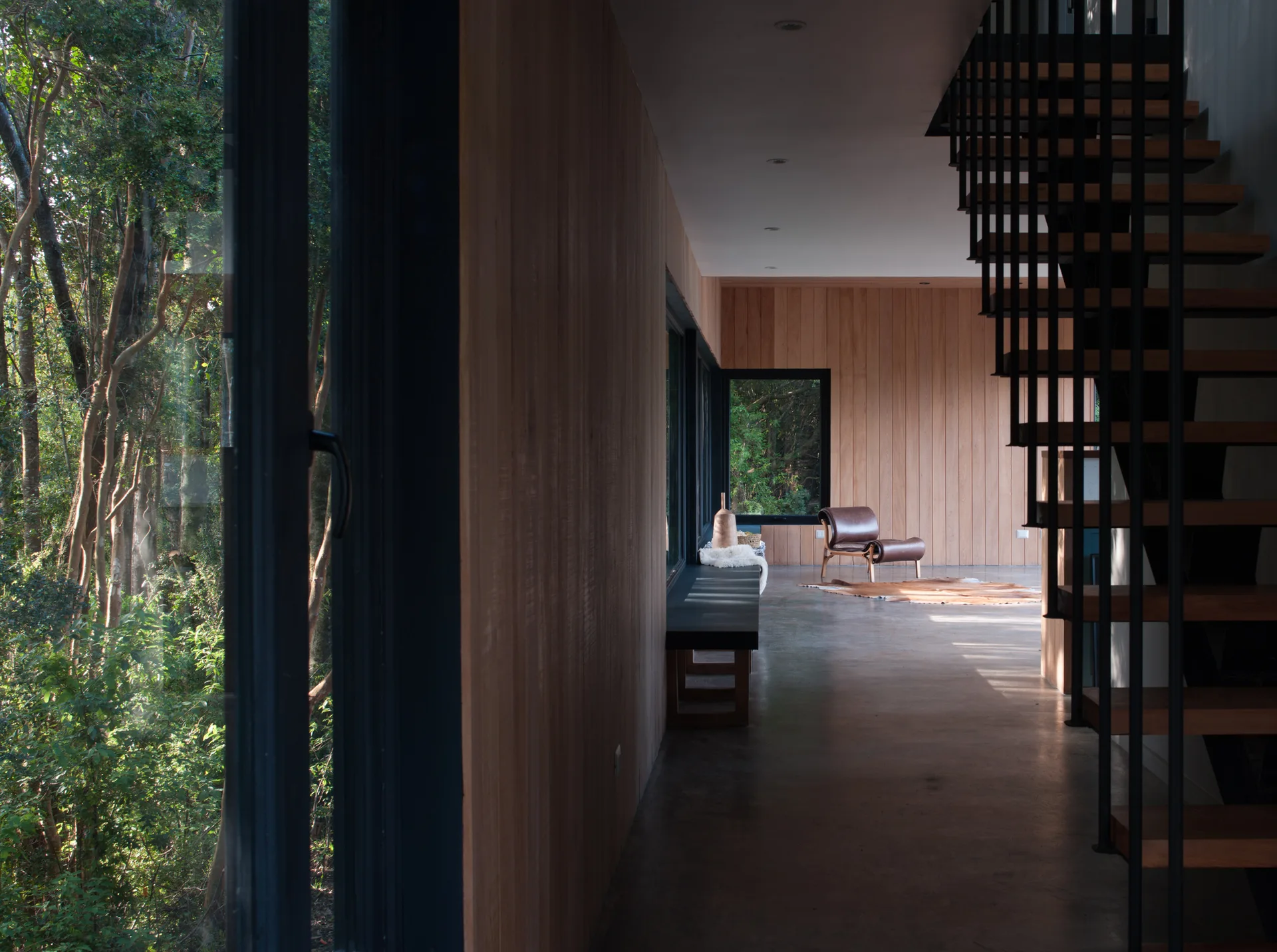 A modern hallway features vertical wood paneling, a gray concrete floor, and an open metal staircase looking out toward a forest.