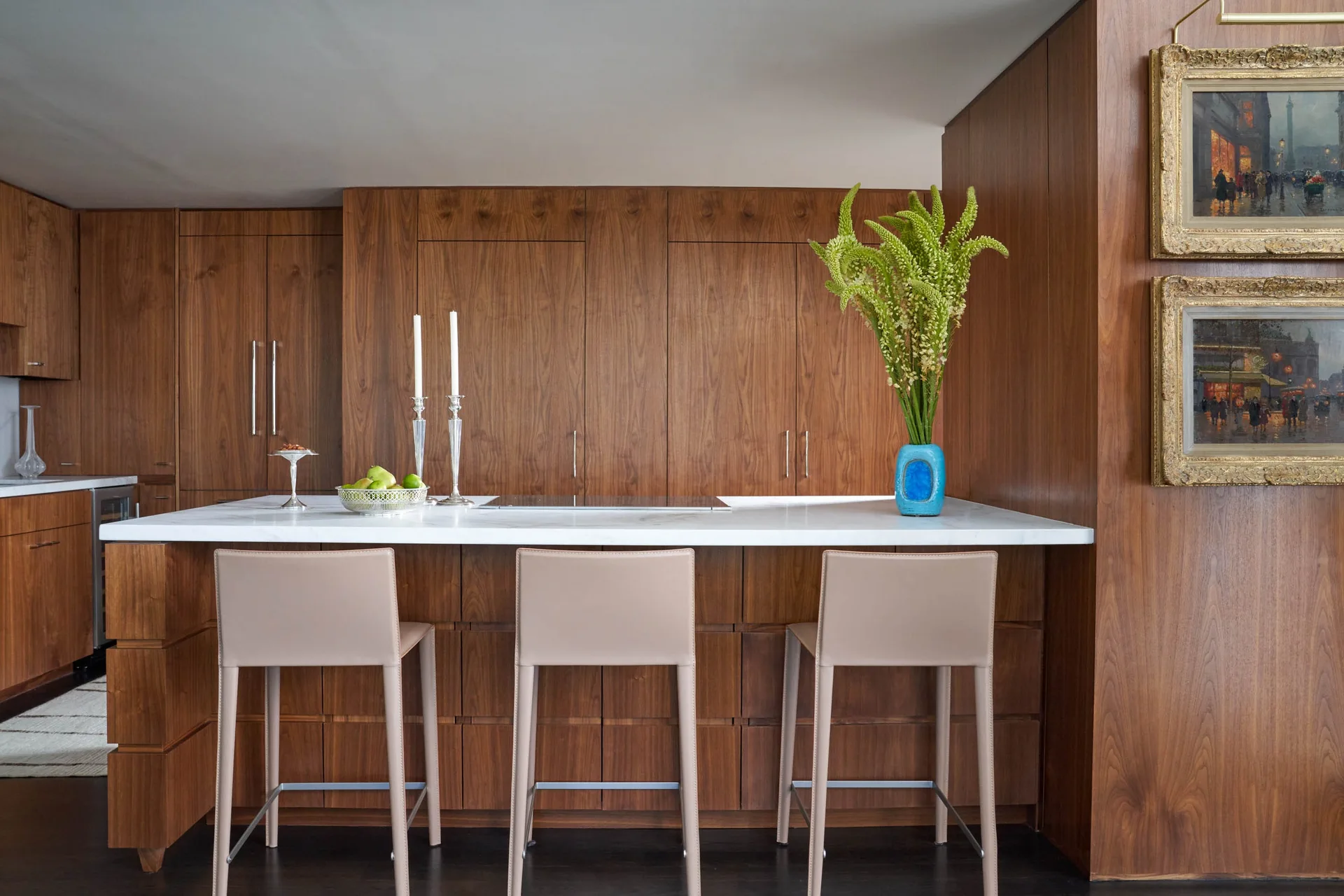 A modern kitchen features floor-to-ceiling walnut cabinets, a white stone island countertop, three beige stools, and a blue accent vase.