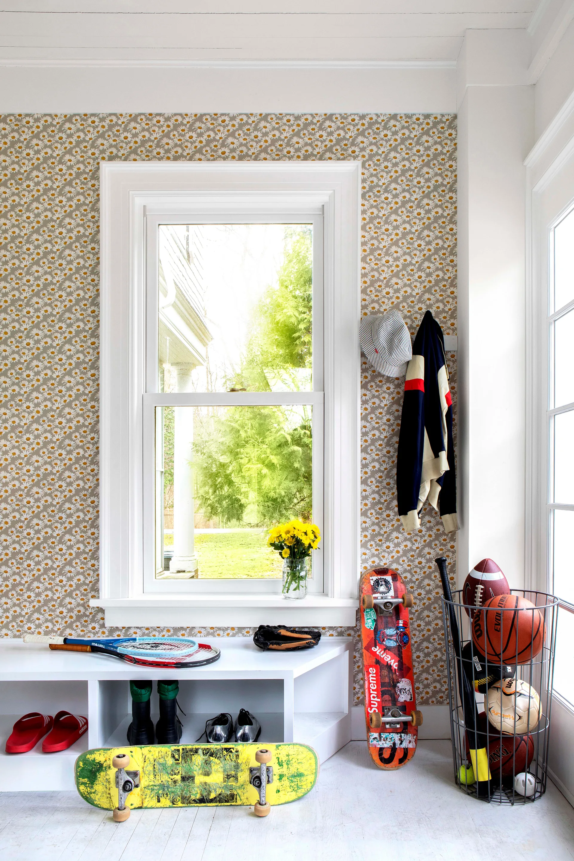 A bright mudroom features daisy patterned wallpaper, a white built-in bench holding sports gear, and a wire basket filled with athletic balls.