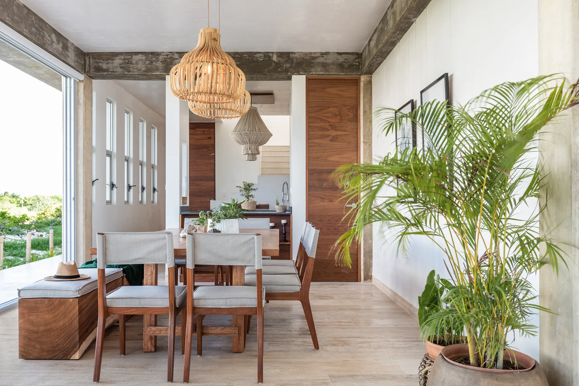 A modern organic dining room featuring a wooden table, light grey chairs, woven pendant lights, and an exposed concrete ceiling beam.