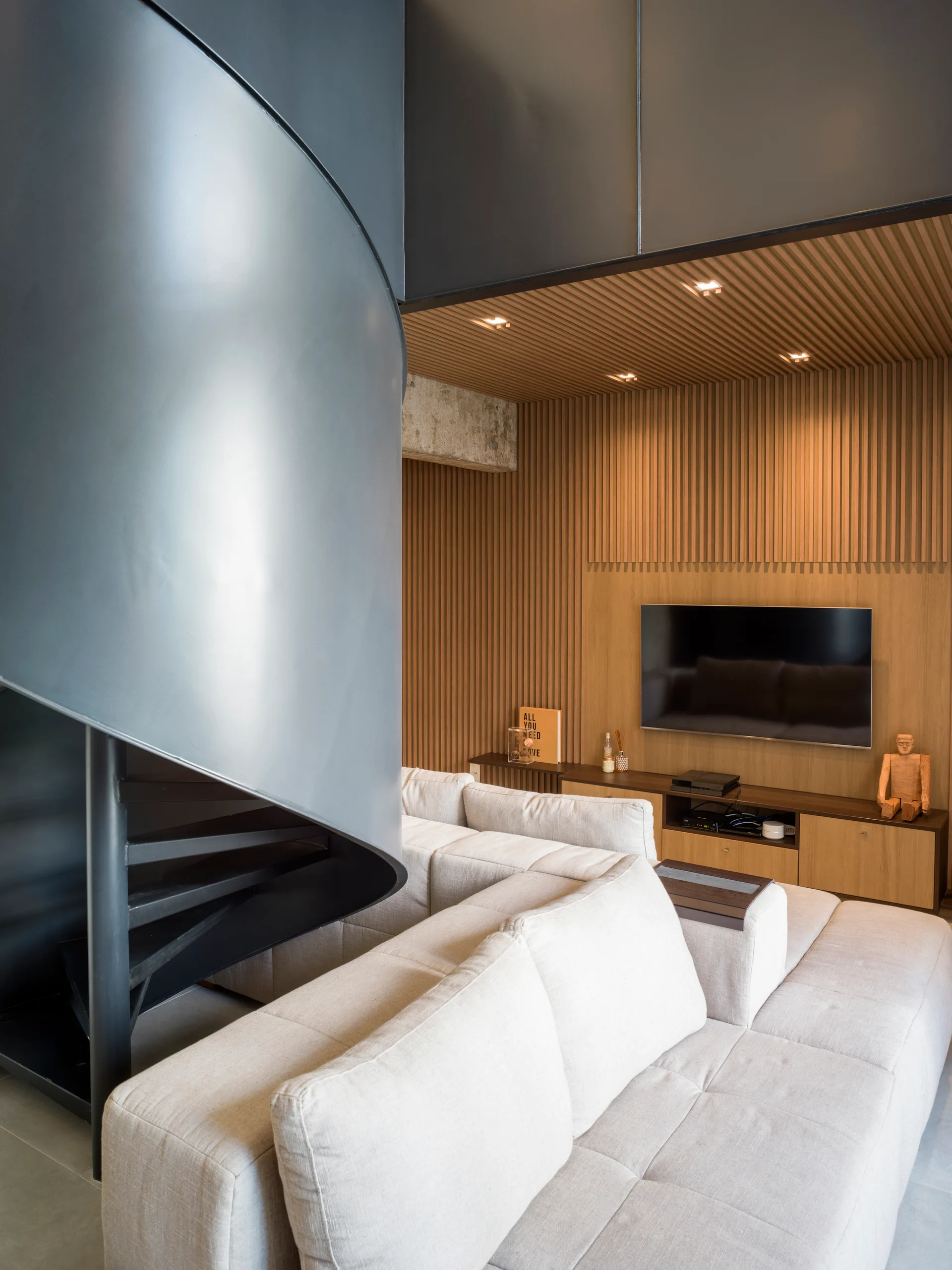 A contemporary living room featuring a beige modular sofa, a black metal spiral staircase, and vertical wood slat paneling on the wall and ceiling.