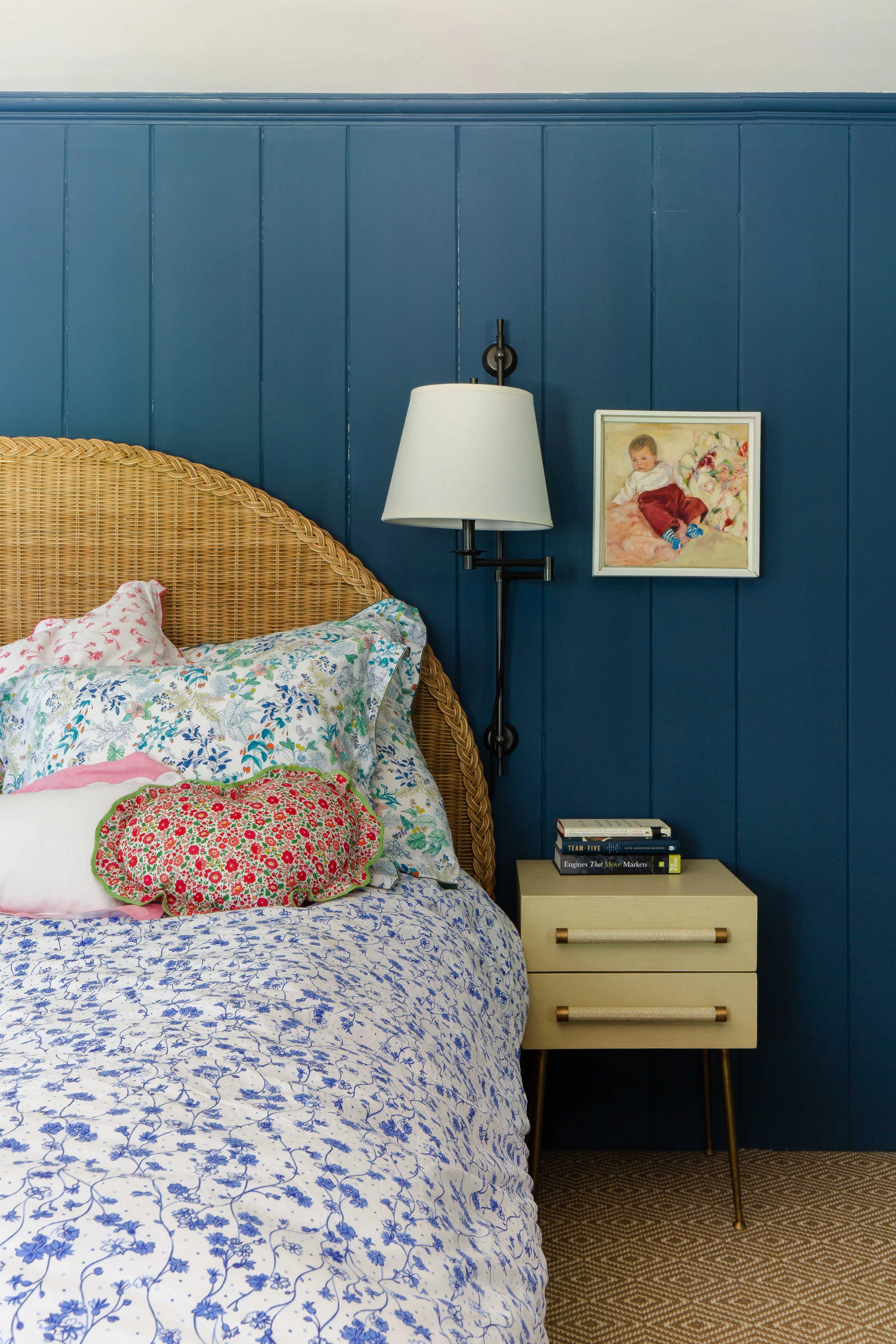 A wicker headboard stands against a dark blue paneled wall next to a light-colored nightstand with books and a wall-mounted brass lamp.