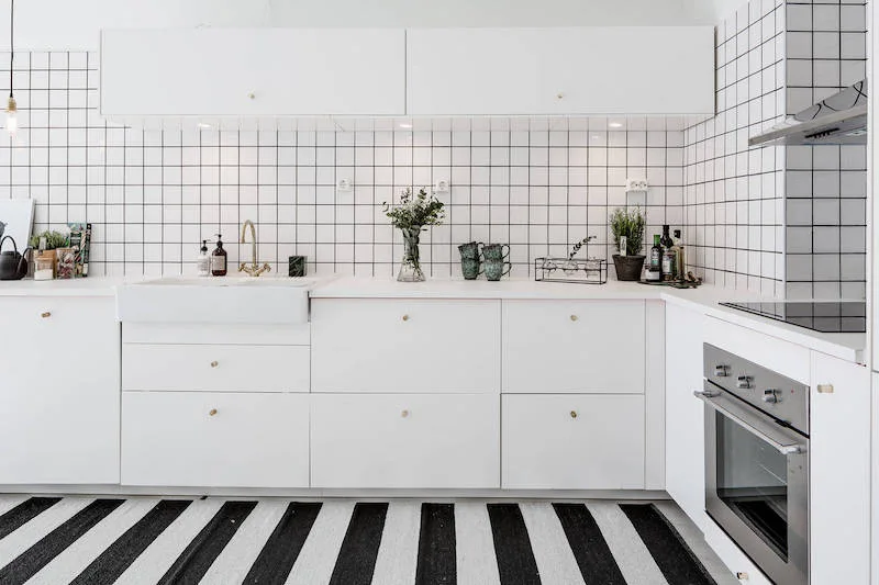 A minimalist white kitchen features flat-front cabinetry, a white grid tiled backsplash with dark grout, and a black and white striped floor runner.