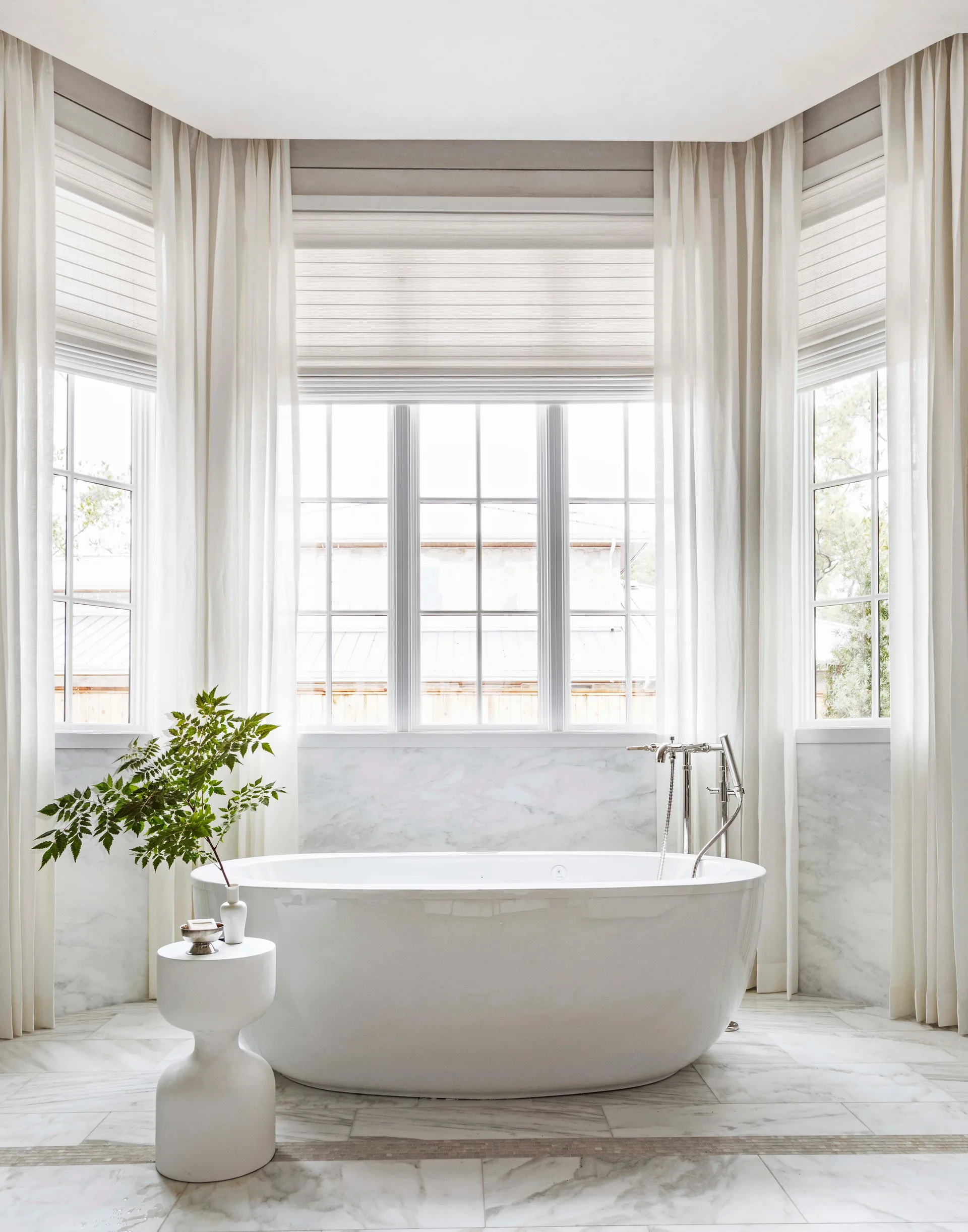 A white freestanding oval bathtub sits in a bright bay window alcove surrounded by marble tile walls, sheer white curtains, and a small side stool.