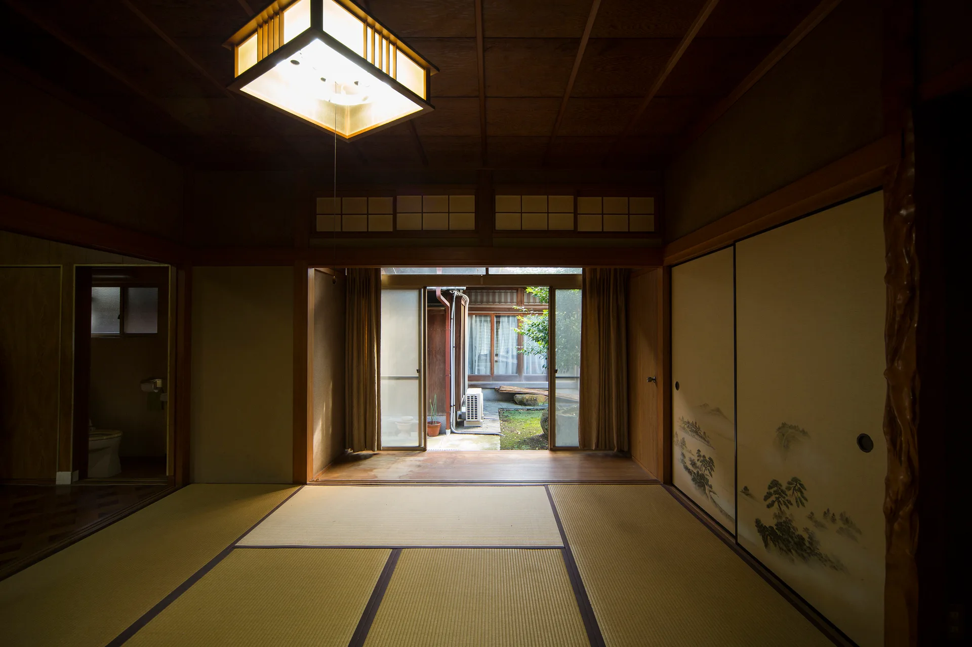 A traditional Japanese room features tatami mats, painted sliding doors, a glowing square ceiling light, and glass doors looking out to a courtyard.