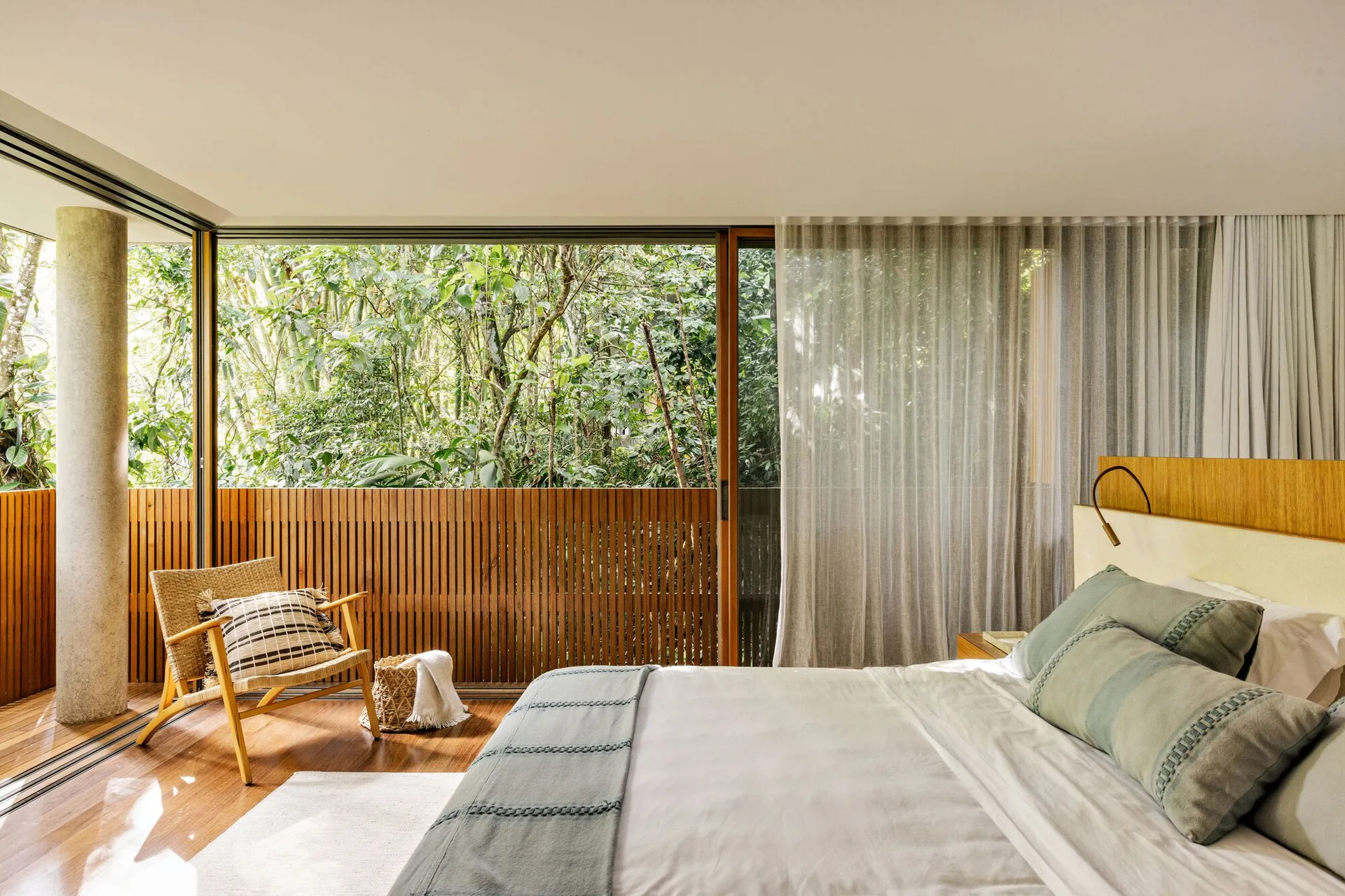 A minimalist bedroom features sage green bedding, a rattan armchair, and sliding glass doors that open to a balcony with a slatted wood screen.