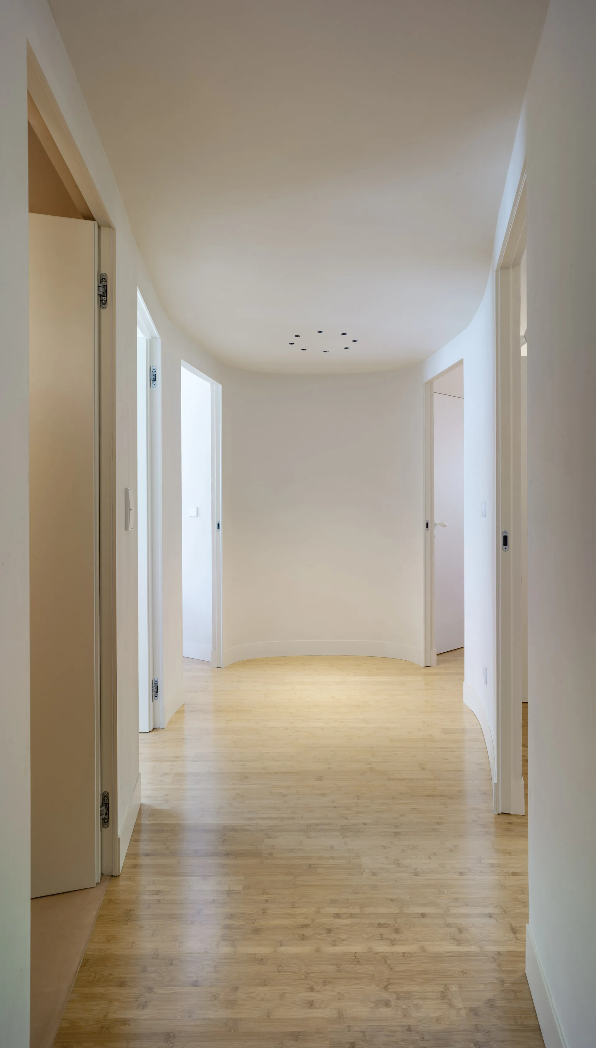A minimalist white hallway features light bamboo wood flooring, multiple open doorways, and a curved far wall.
