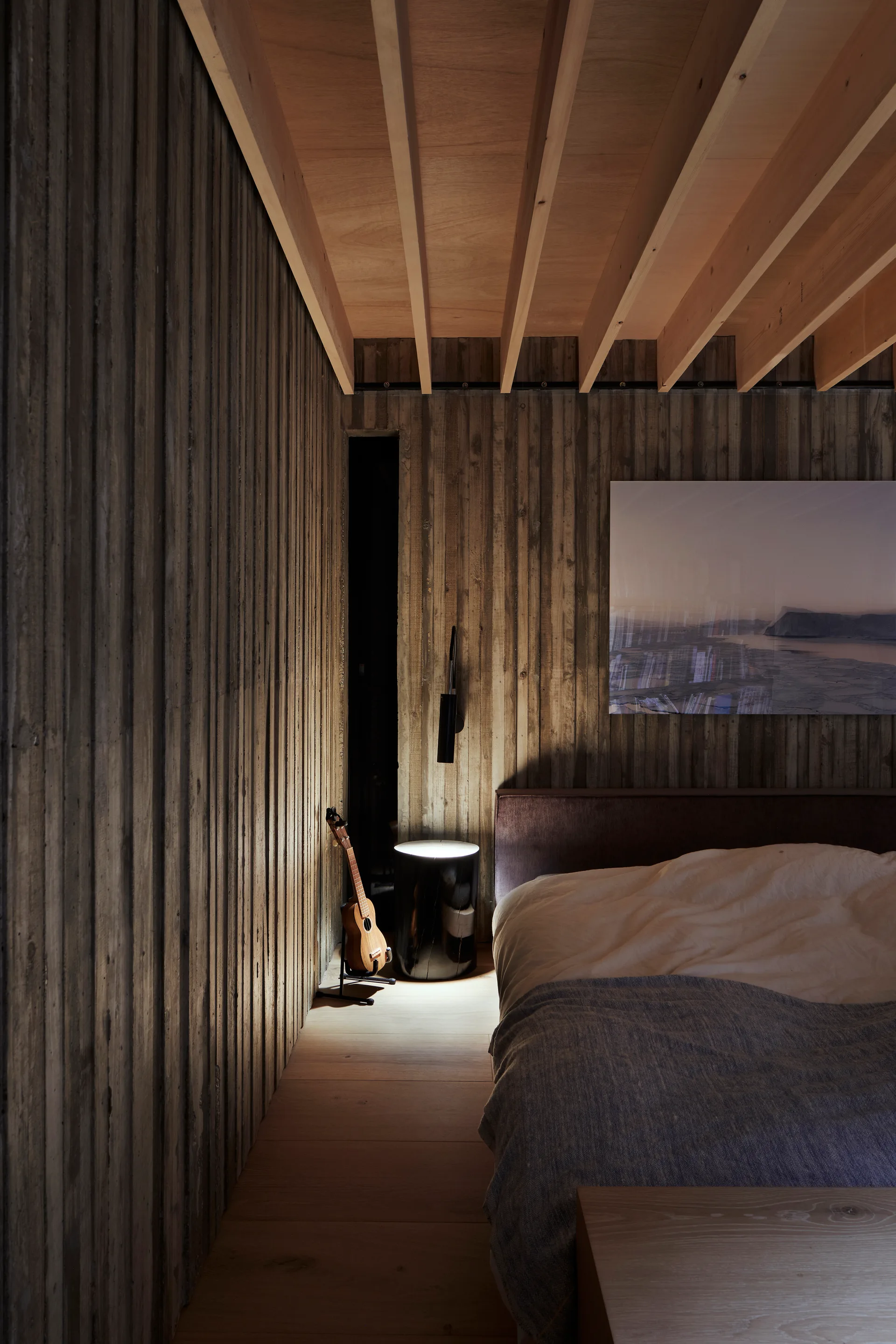 A minimalist bedroom featuring vertical board-formed concrete walls, an exposed light wood ceiling, a partially visible bed, and a dark side table.