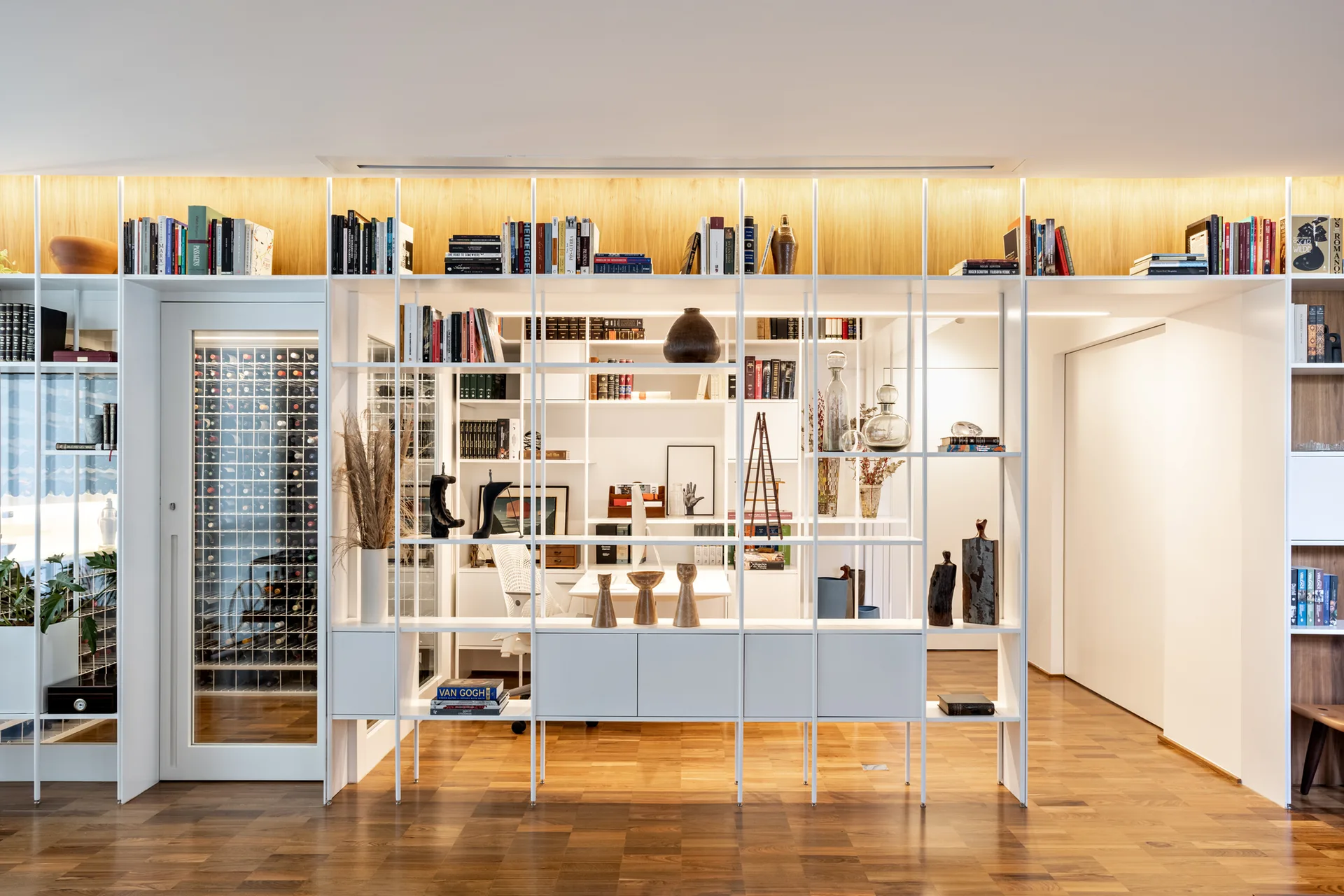 A large white grid shelving unit acts as a room divider, displaying books and art while framing a home office desk and a glass-enclosed wine cellar.
