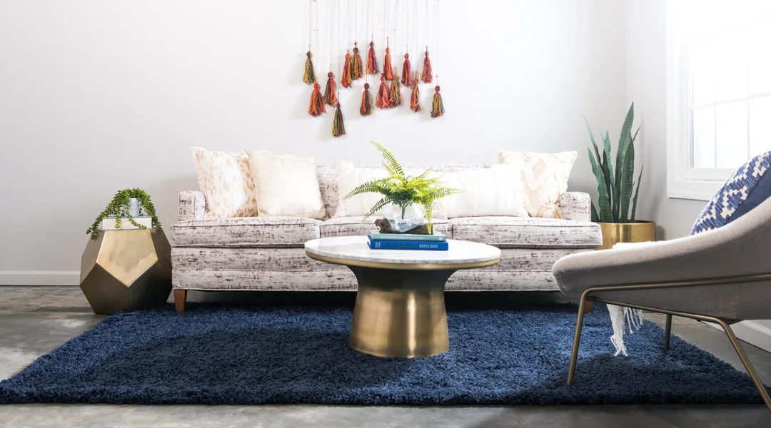 A living room with a patterned sofa, a round brass coffee table with a marble top, a geometric gold side table, and a plush navy blue shag rug.