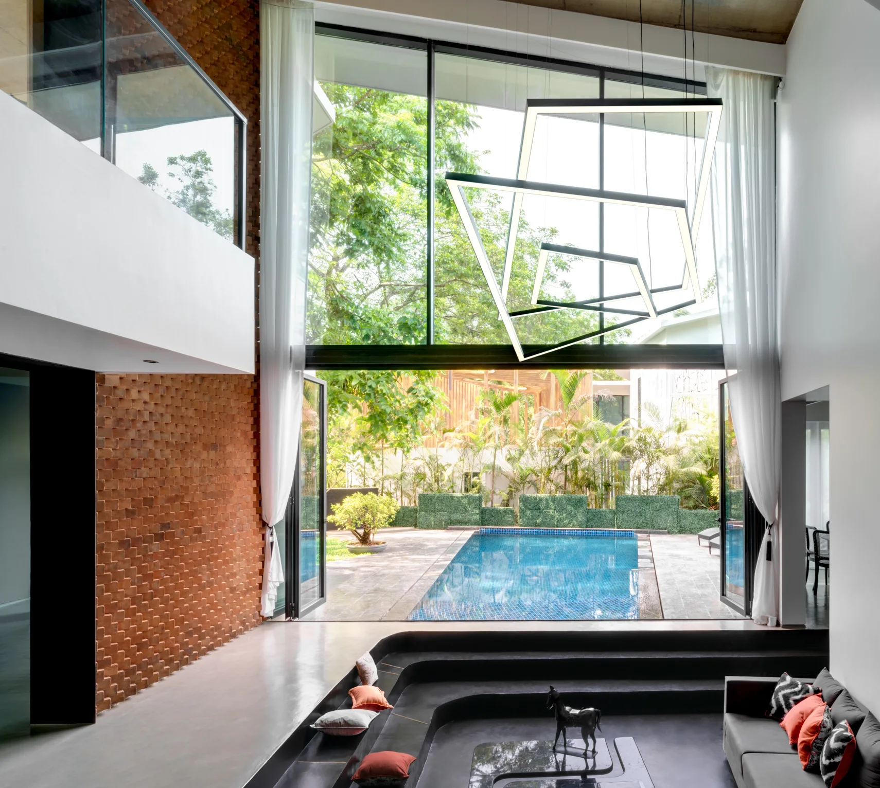 A modern sunken living area features a textured brick accent wall, geometric hanging lights, and floor-to-ceiling windows showing a backyard pool.
