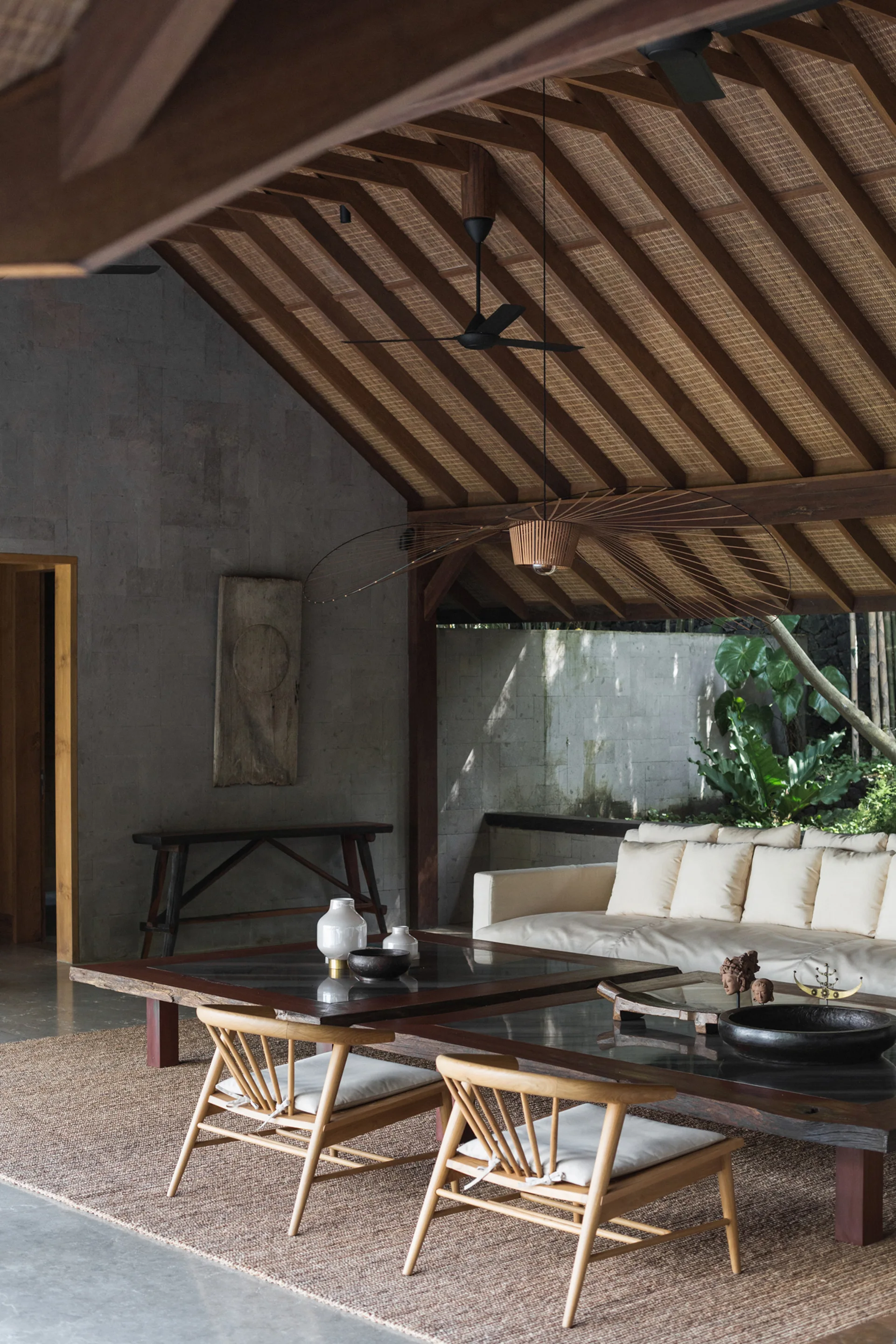 An open-air pavilion living room features a cream sofa, low wood coffee tables, and wooden chairs on a woven rug under a vaulted wood ceiling.
