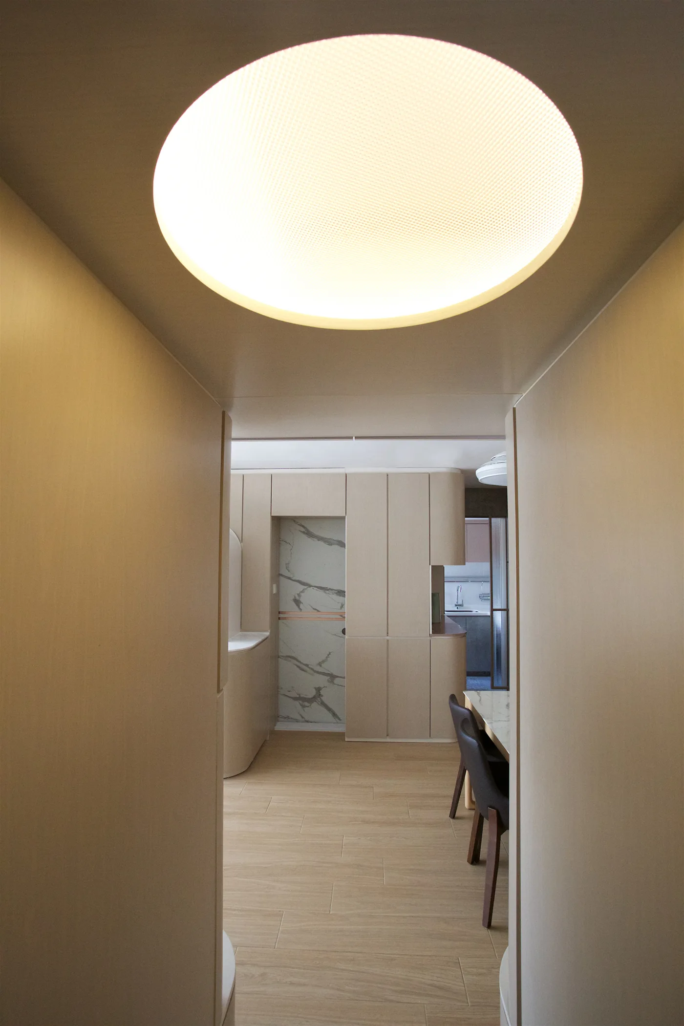 A minimalist hallway lined in light wood paneling with a large circular recessed light overhead looking toward a kitchen and dining area.