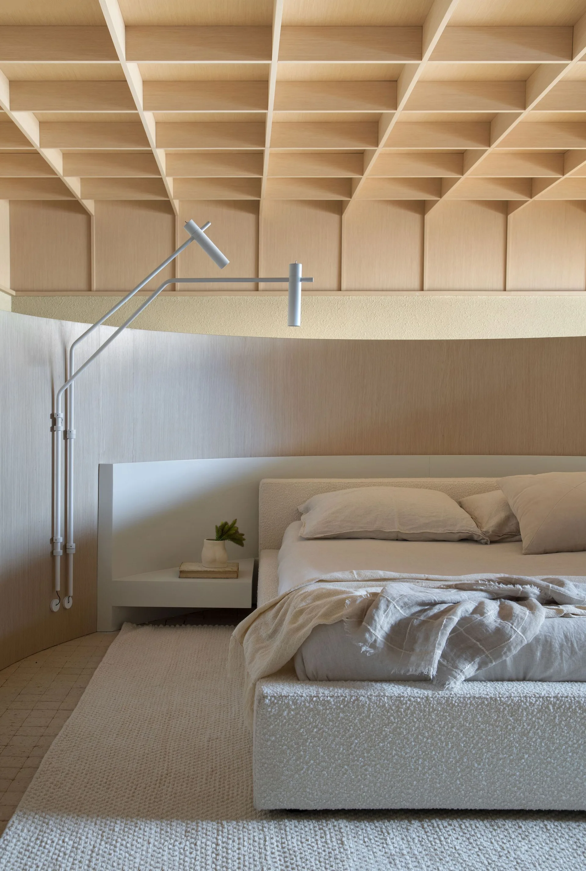 A neutral bedroom features a boucle upholstered bed against a curved wood-paneled wall below a light oak coffered grid ceiling.