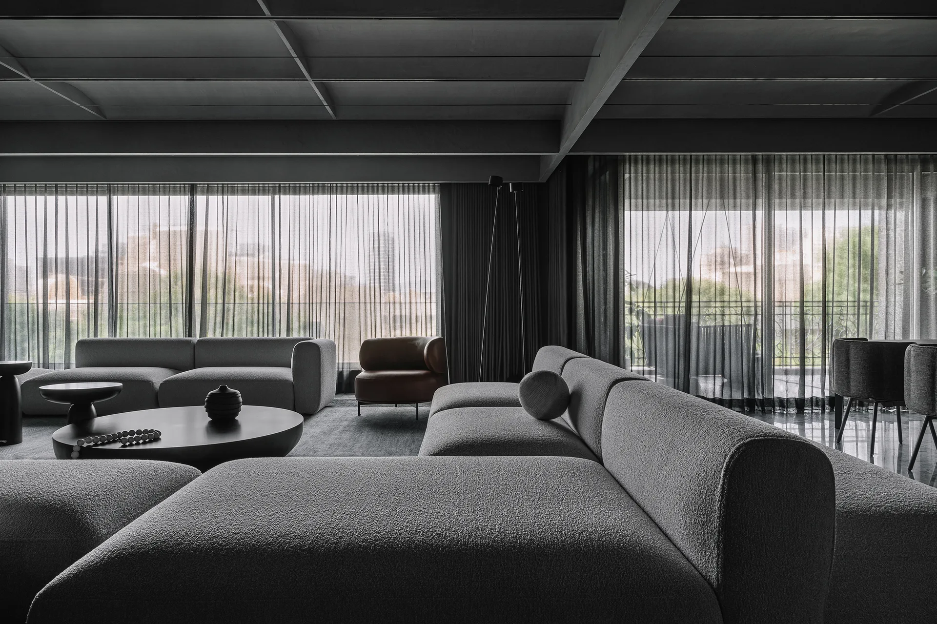 A spacious dark grey living room with low-profile textured sectional sofas, a round black coffee table, and sheer floor-to-ceiling curtains.