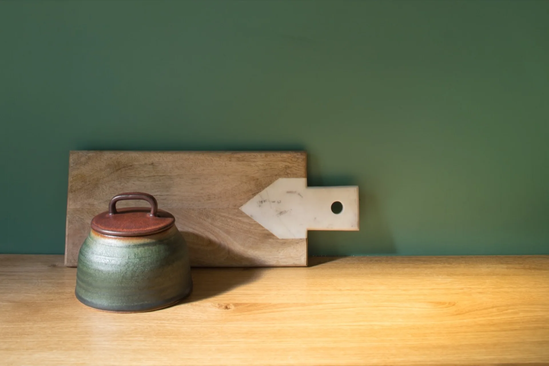 A lidded green ceramic jar and a wooden cutting board with white marble inlay rest on a wood countertop against a solid green wall.