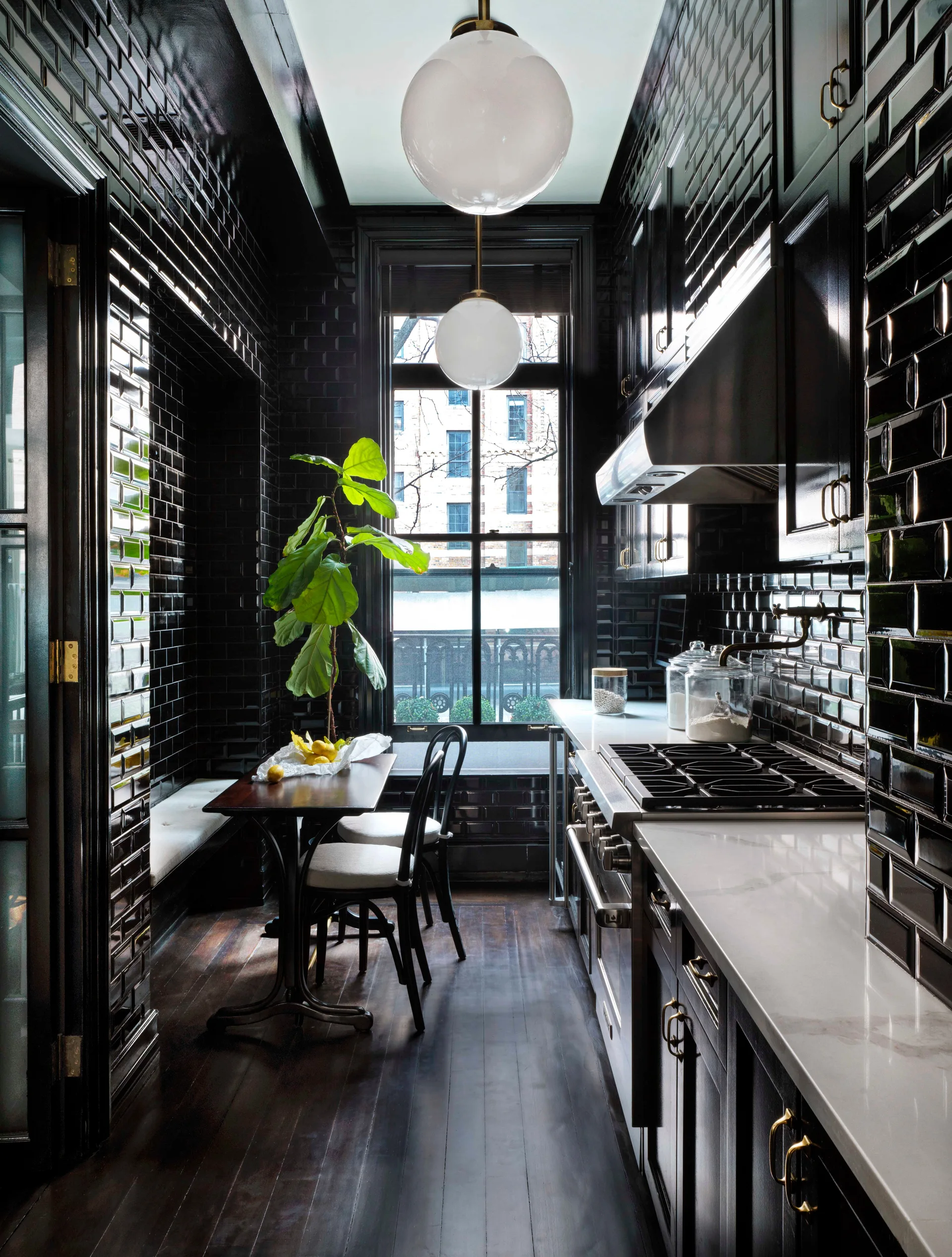 A narrow galley kitchen features reflective black subway tile walls, matching black cabinets, white countertops, and a small dining nook with a built-in bench.