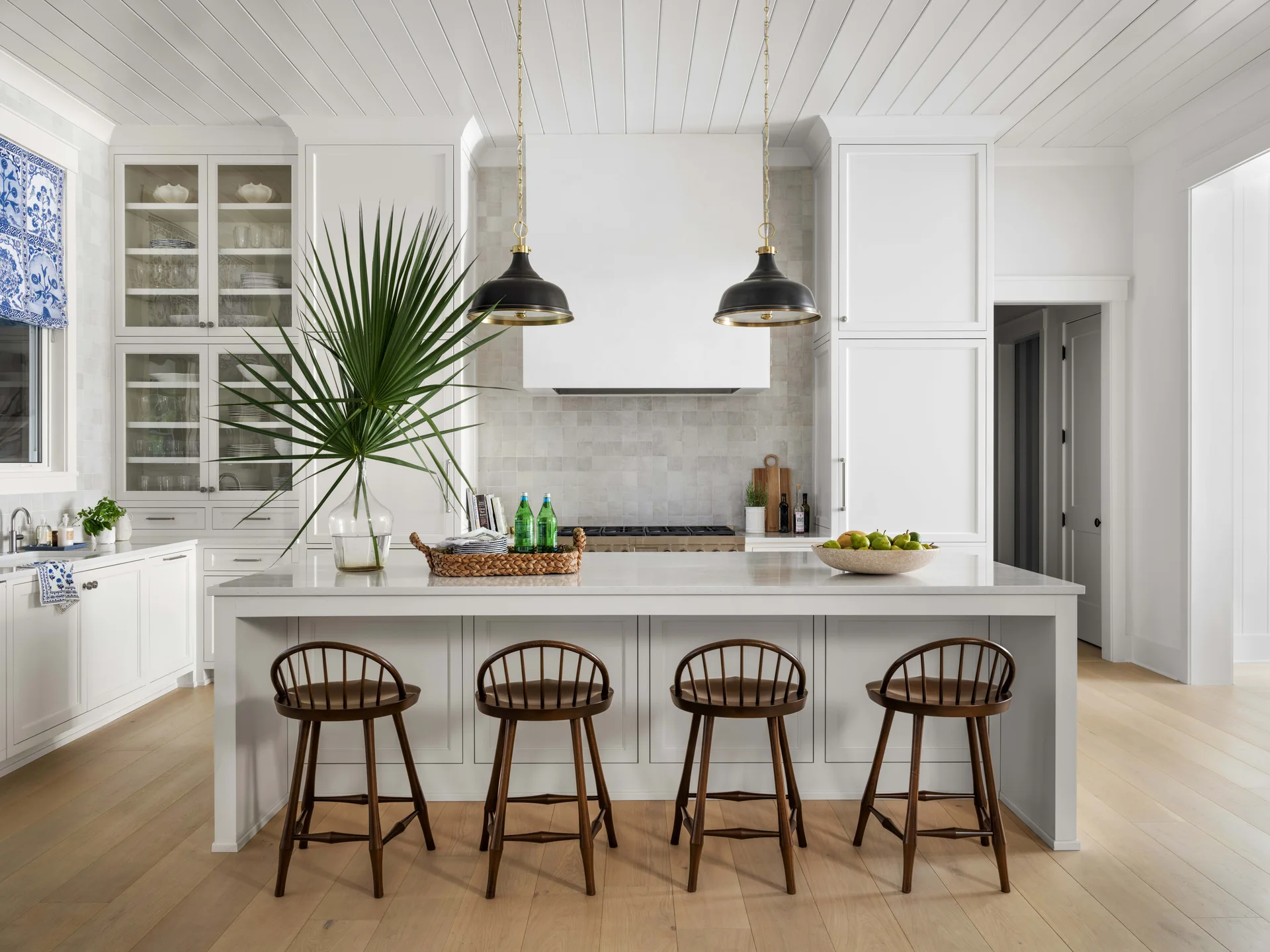 Four wooden bar stools sit at a white kitchen island under two black pendant lights in a bright kitchen with white cabinetry.