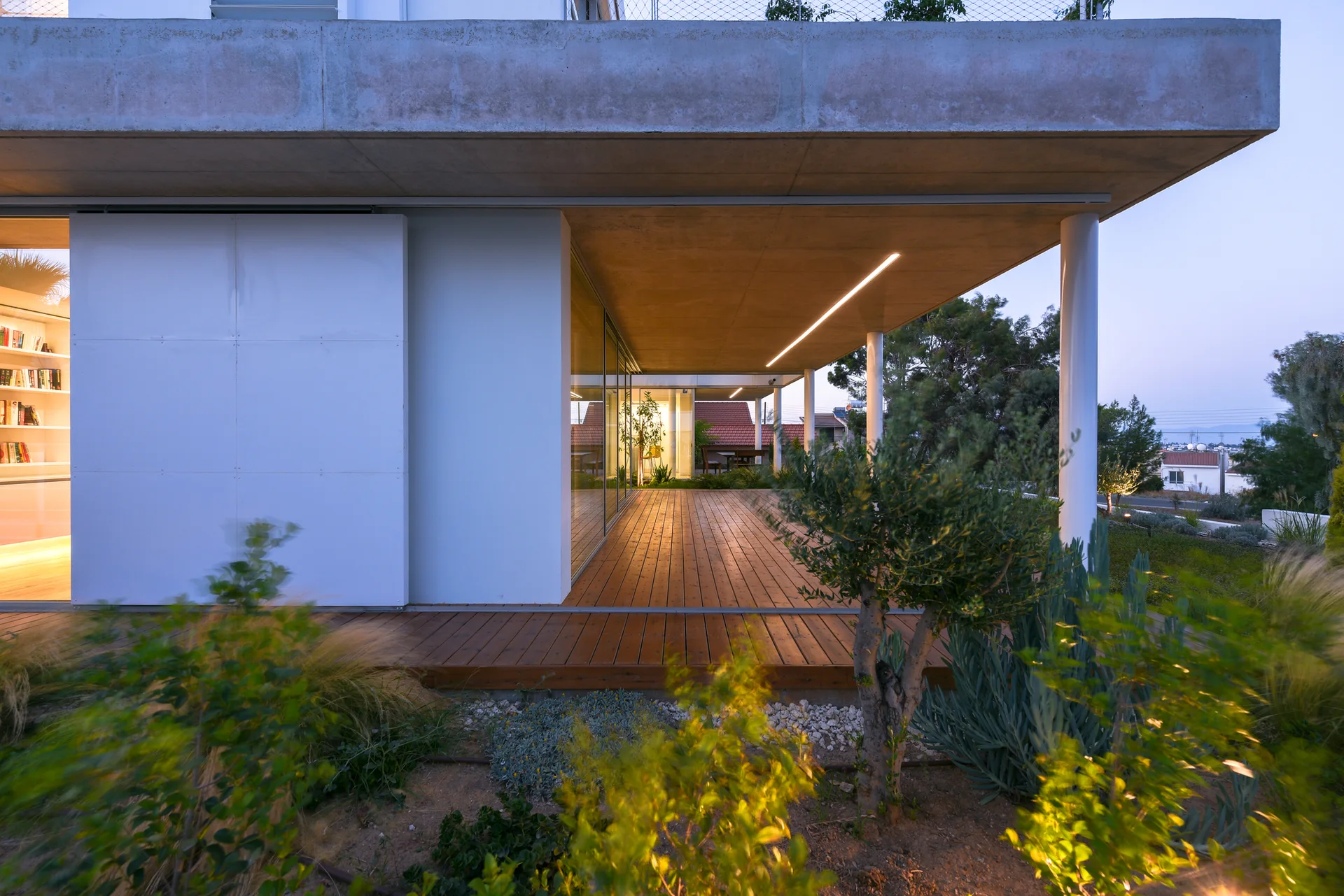 A modern residence features a covered wooden deck beneath a concrete canopy next to floor-to-ceiling windows and a landscaped garden.