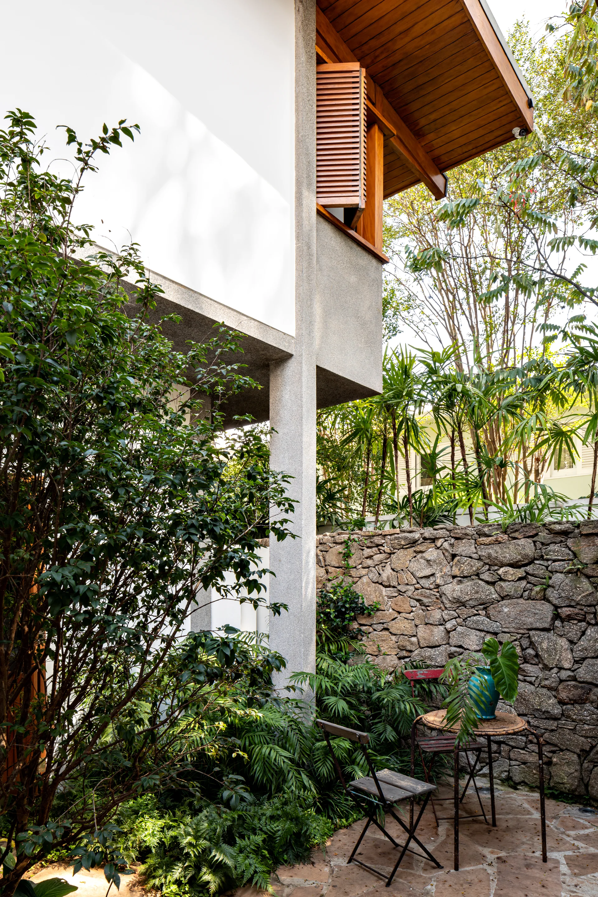 A small garden patio features a metal bistro table and chairs next to a rustic stone retaining wall below a modern house exterior with wood shutters.