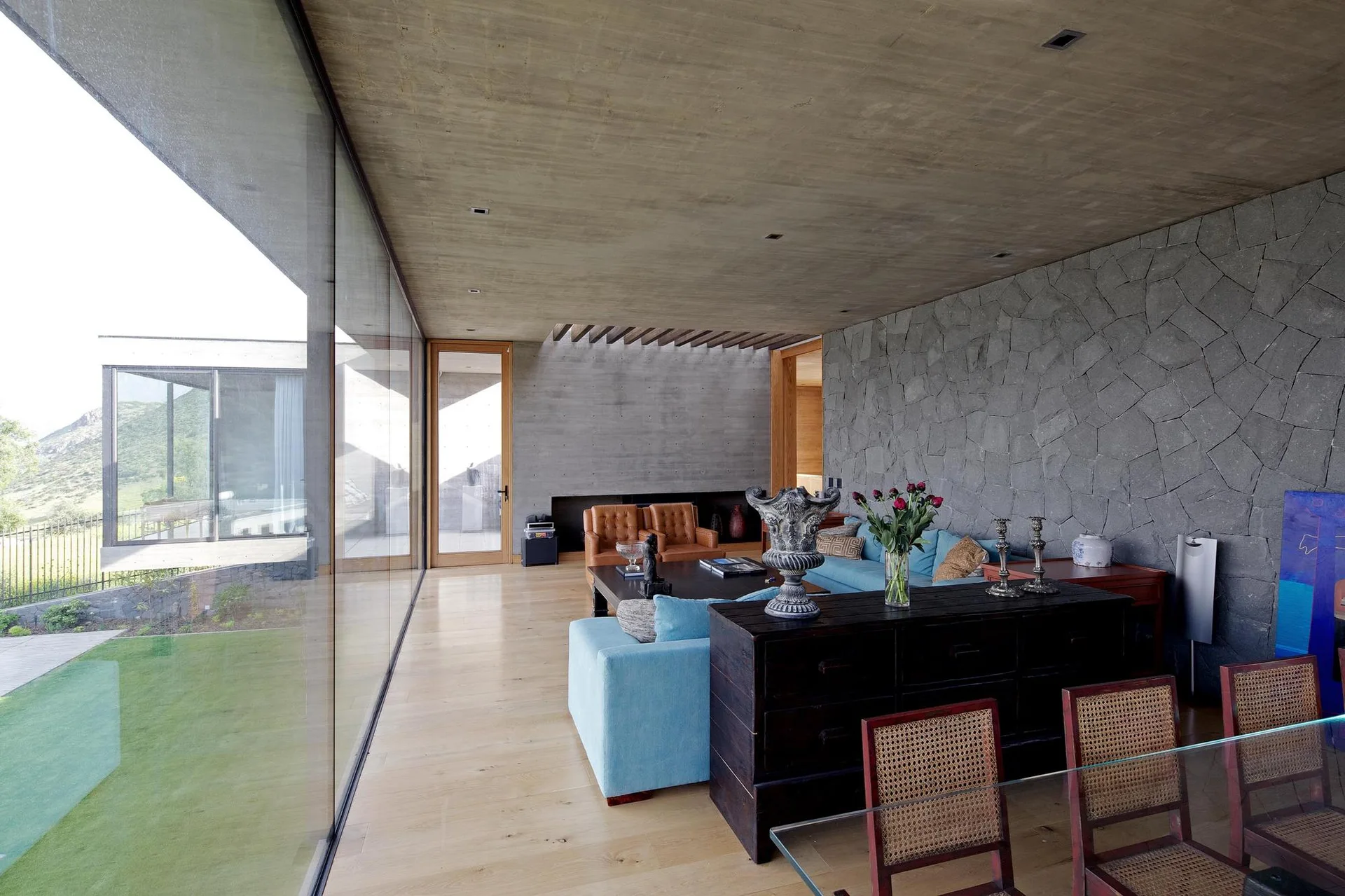 A modern living room featuring a floor-to-ceiling glass wall, an exposed concrete ceiling, a gray stone accent wall, blue sofas, and light wood floors.