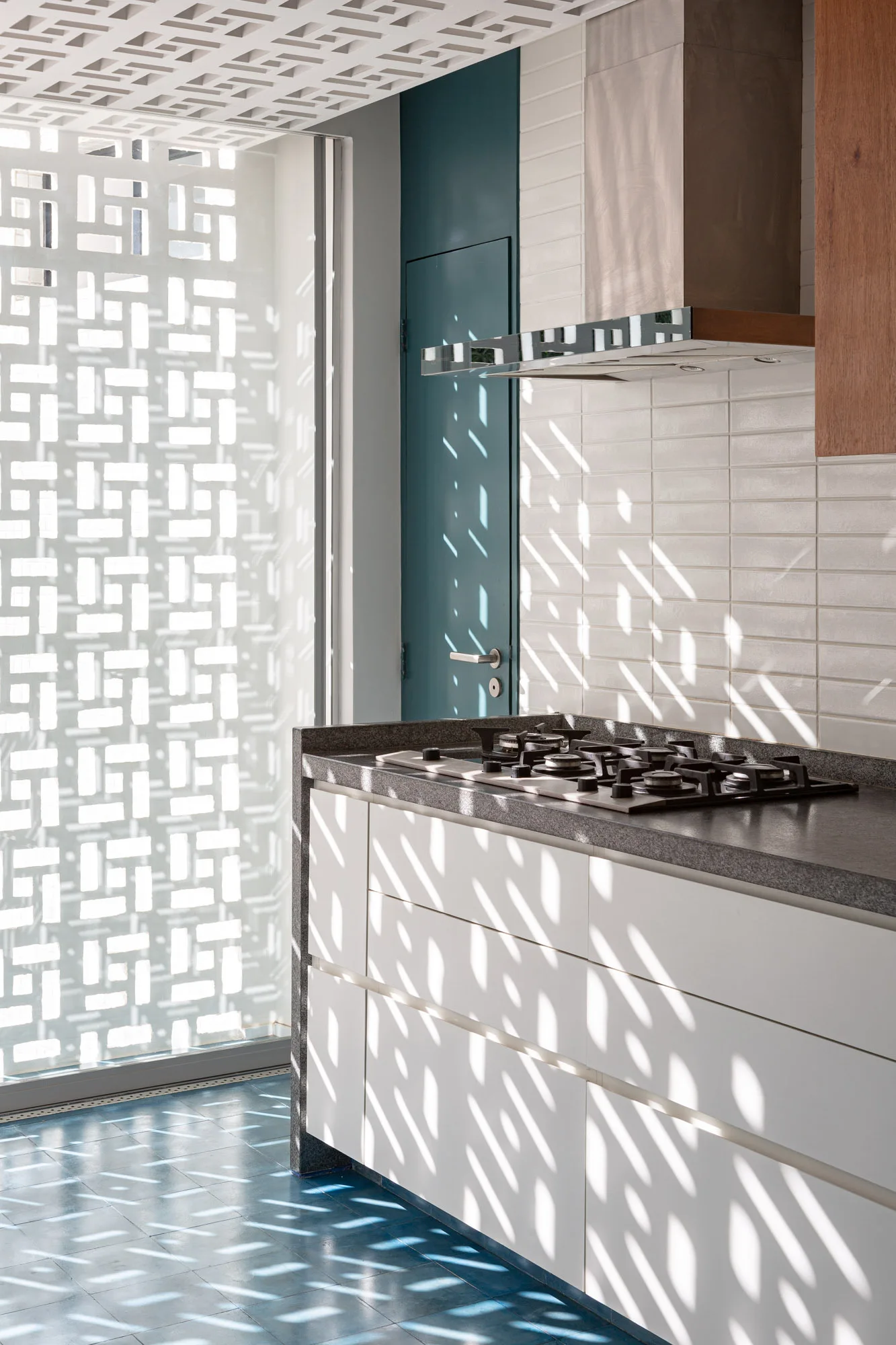 A white kitchen counter with a gas stove stands next to a breeze block screen that filters sunlight across blue floor tiles.