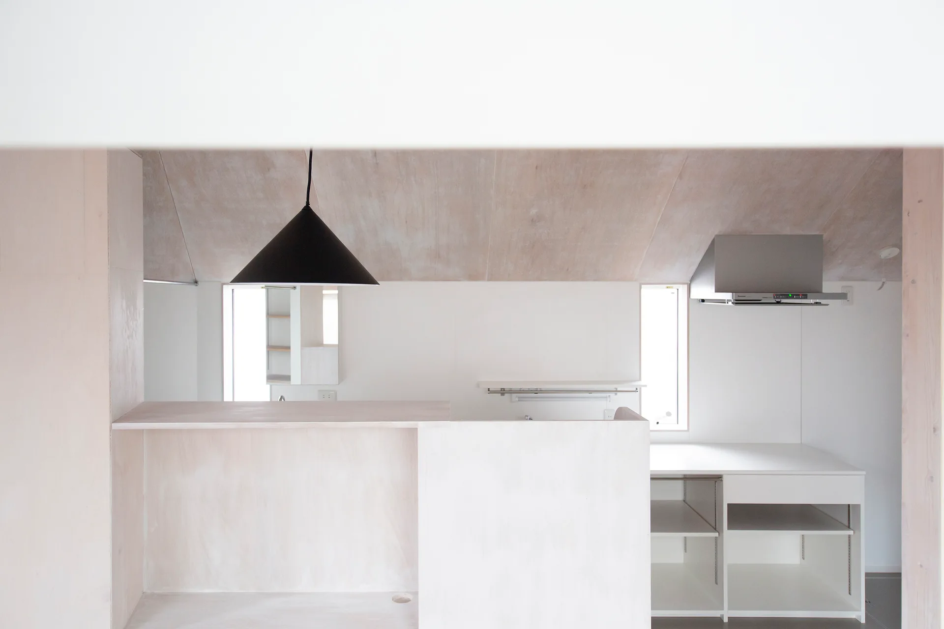 A minimalist kitchen features white walls, a sloped plywood ceiling, a black cone pendant light, and light wood counters under two narrow windows.