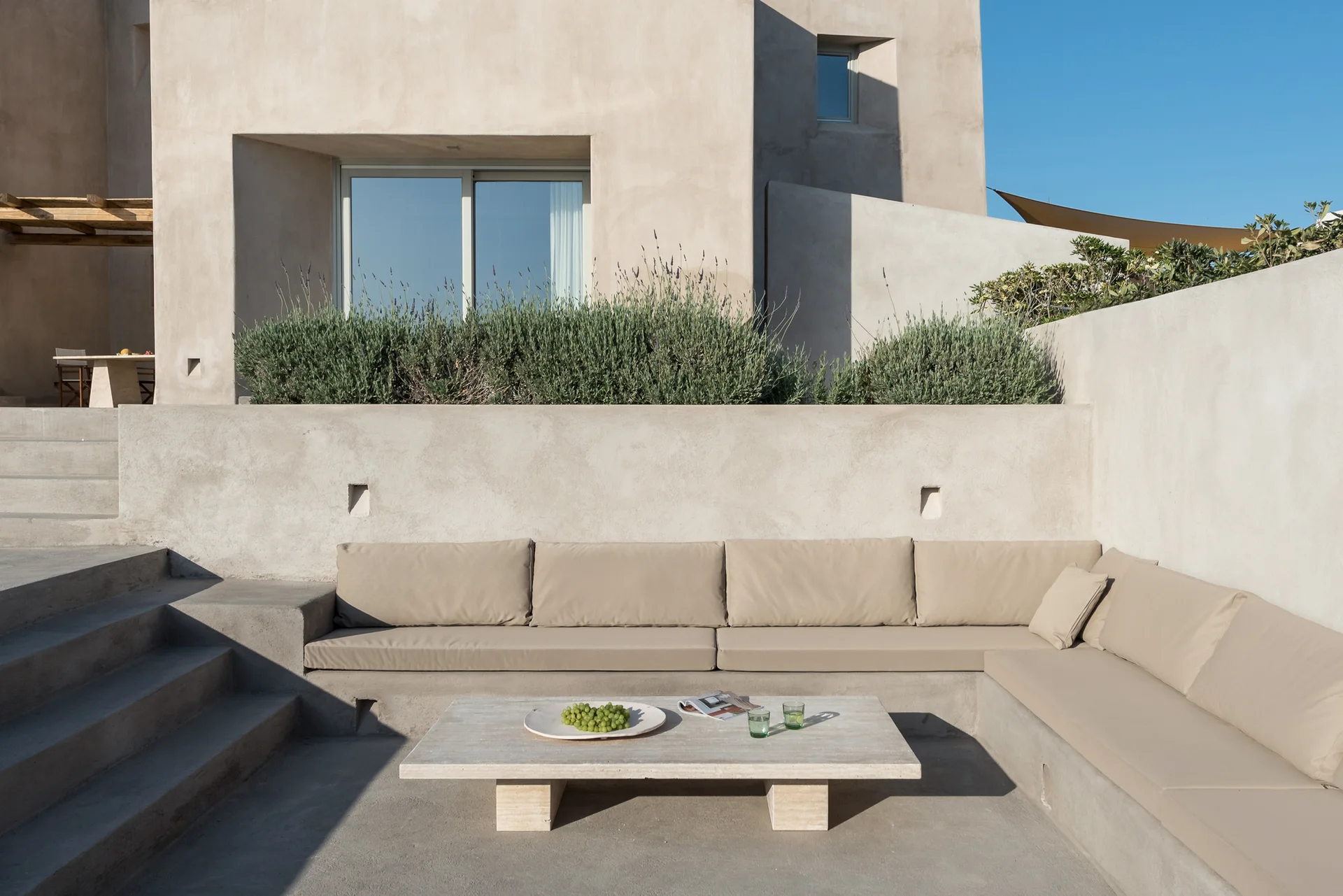 A minimalist concrete patio with an L-shaped built-in sofa, tan cushions, and a low stone coffee table in front of a stucco building.