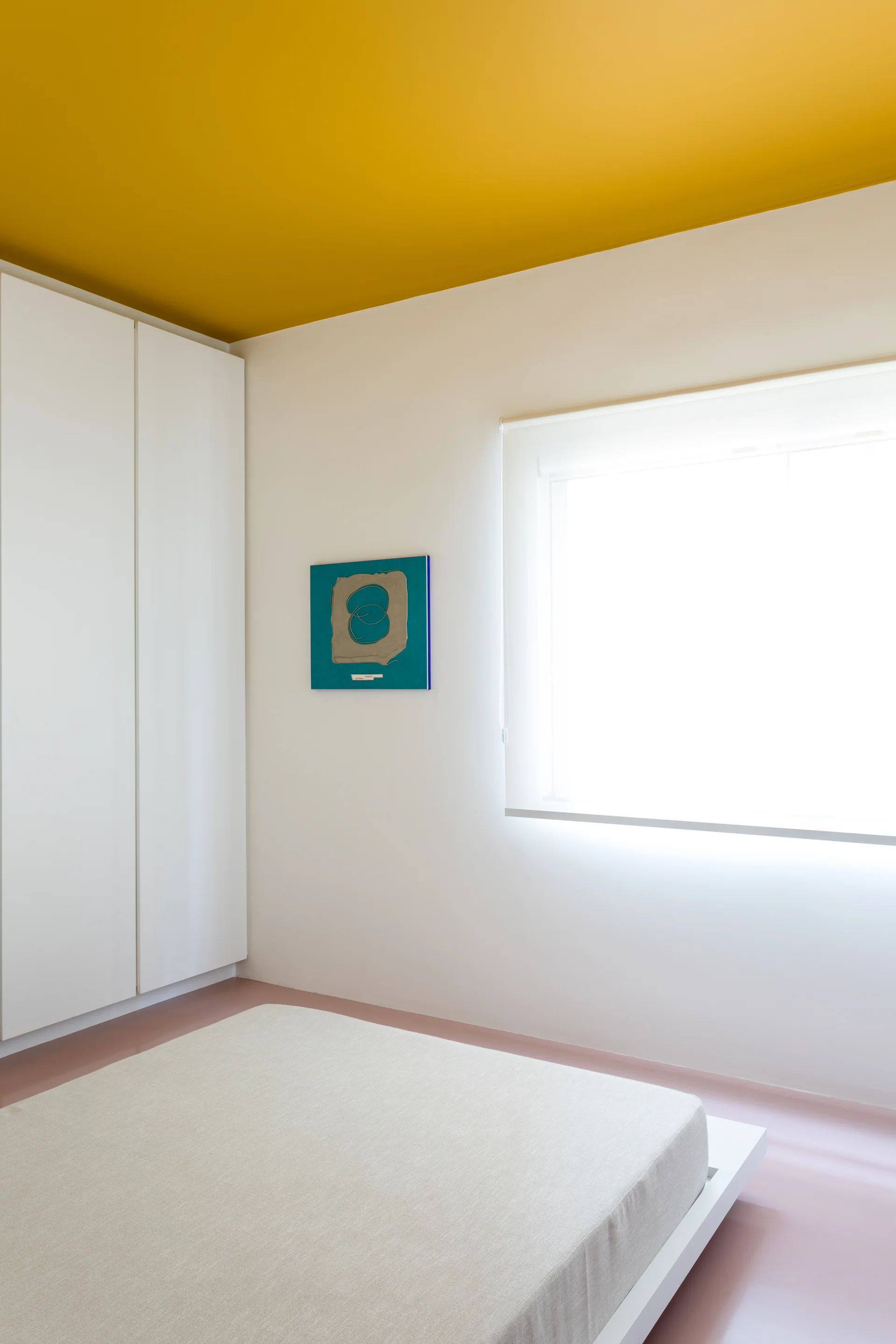 A minimalist bedroom features a low platform bed, a white built-in wardrobe, a bright yellow ceiling, and a polished pink floor.