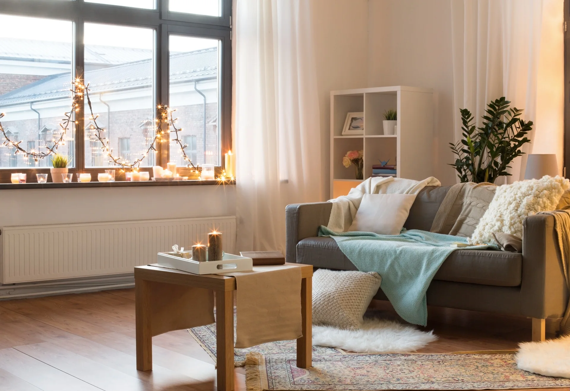 A grey sofa with layered throw blankets and pillows sits near a window decorated with glowing string lights and candles in a living room.