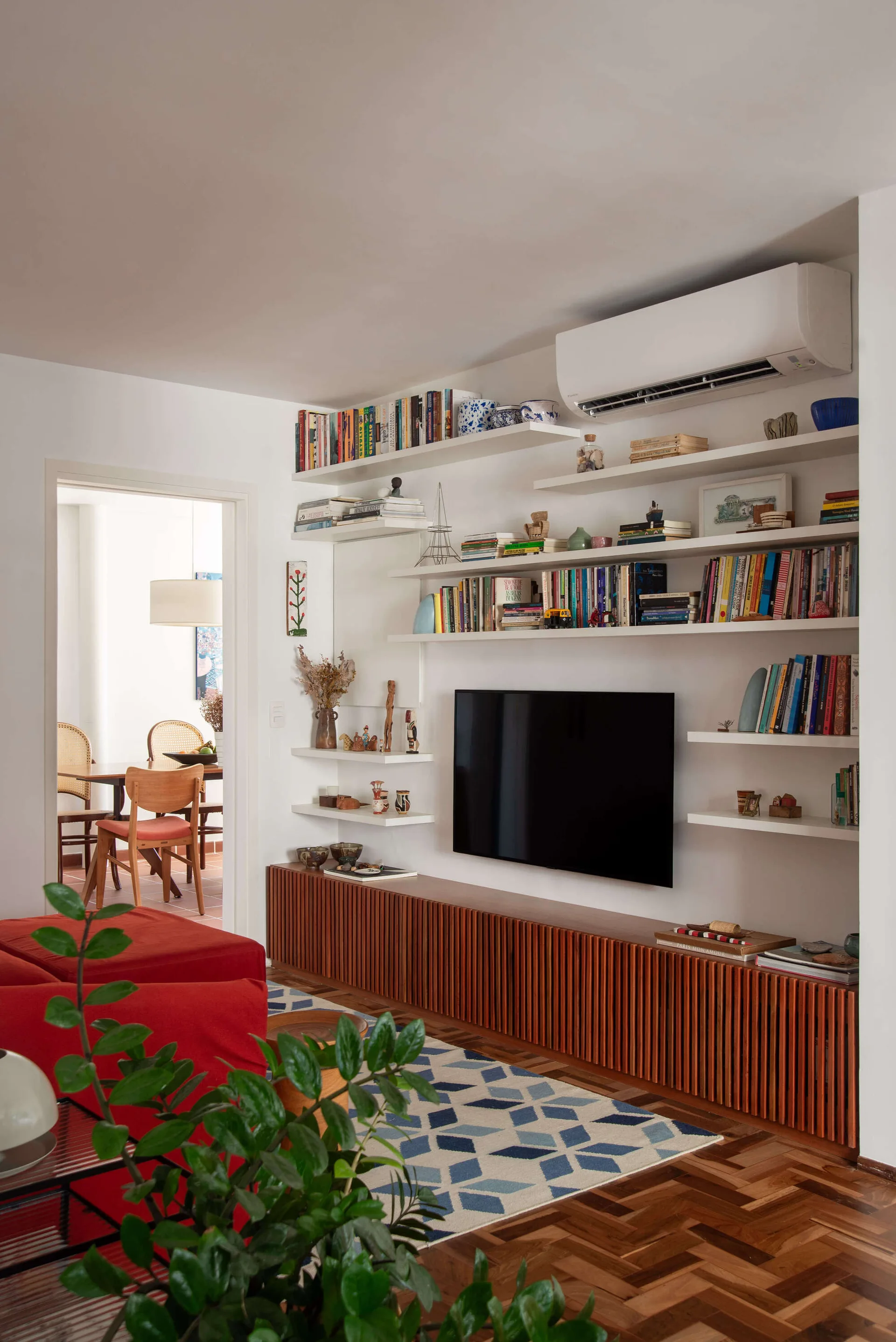 A living room features a wall-mounted TV surrounded by white floating shelves above a slatted wood console and a red sofa.