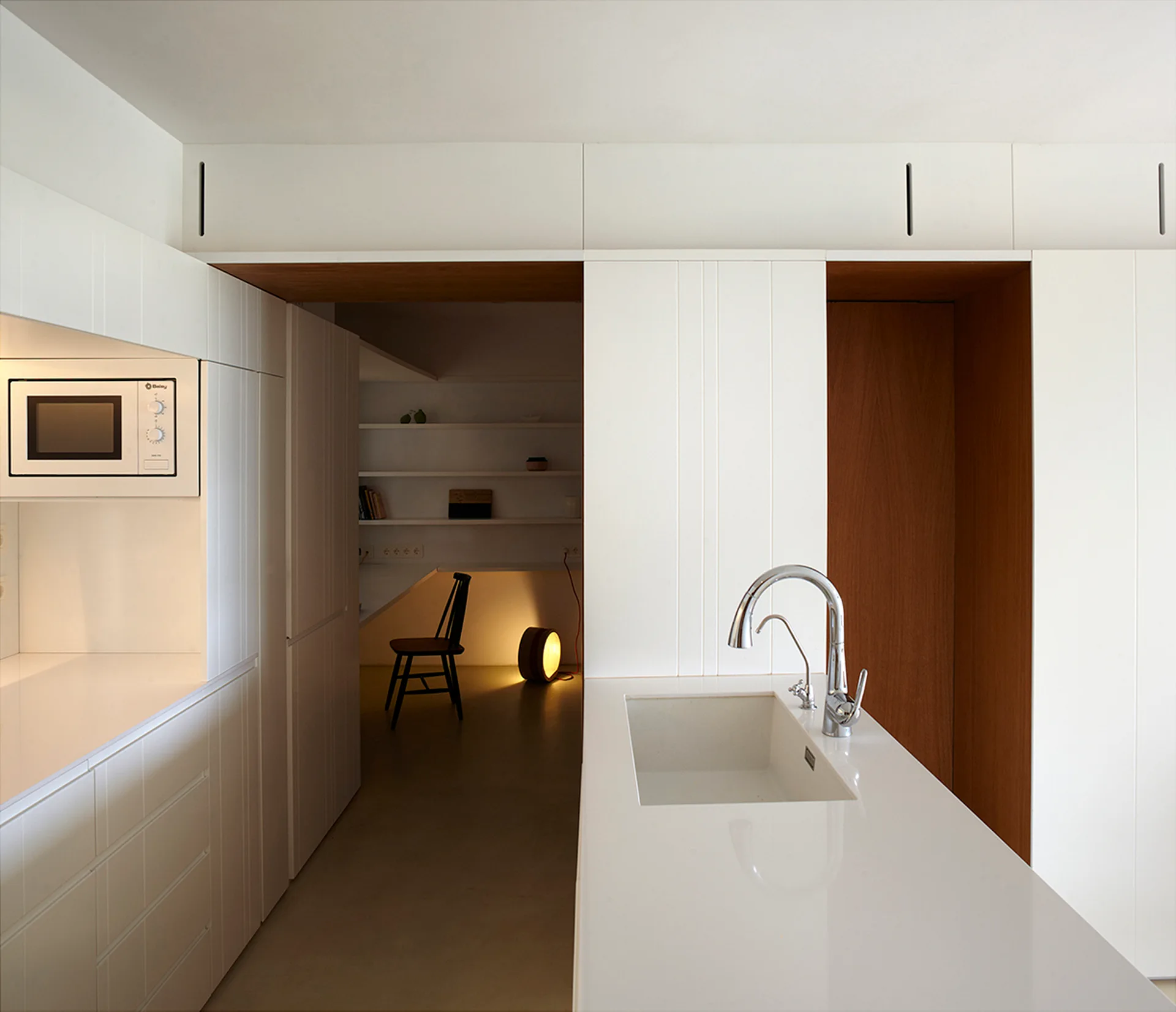 A minimalist white kitchen island with a chrome faucet overlooks a warm, wood-framed entryway leading to an adjacent study nook with a black chair.
