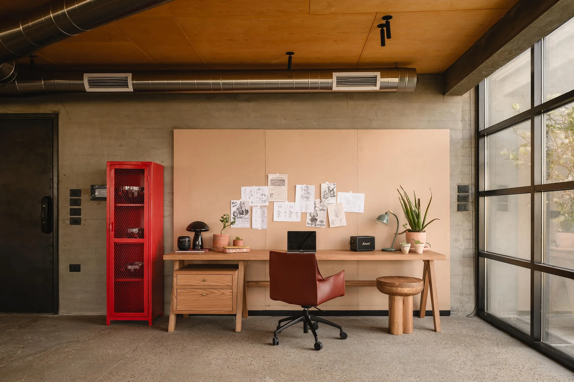 An industrial home office features a long wooden desk with a laptop, a red leather chair, a bright red tall metal cabinet, and exposed concrete walls.