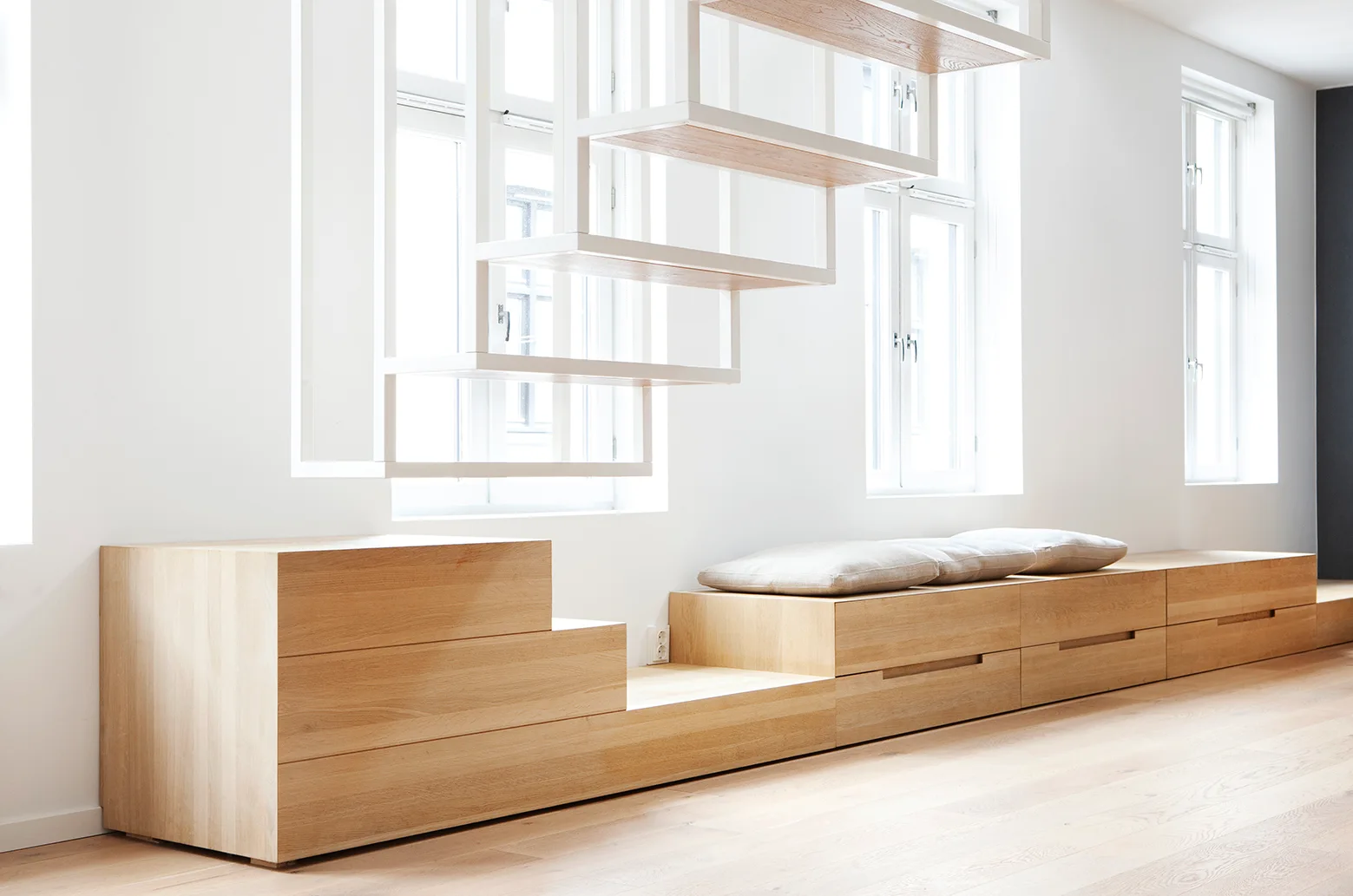 A minimalist white room features a long, low light-wood storage bench with grey cushions beneath a floating wooden staircase by large windows.