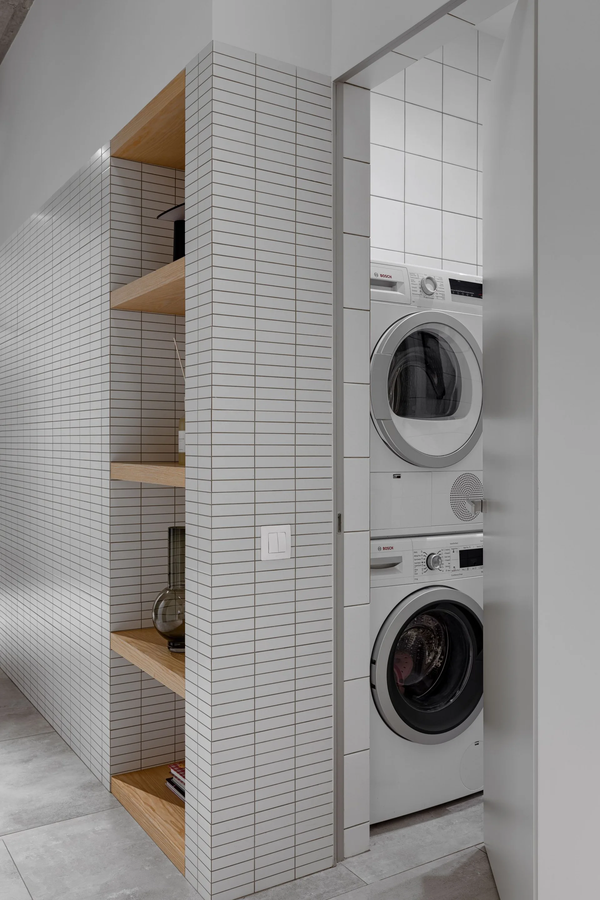 A white tiled partition wall features built-in wood shelves next to a doorway revealing a stacked washer and dryer in a laundry alcove.