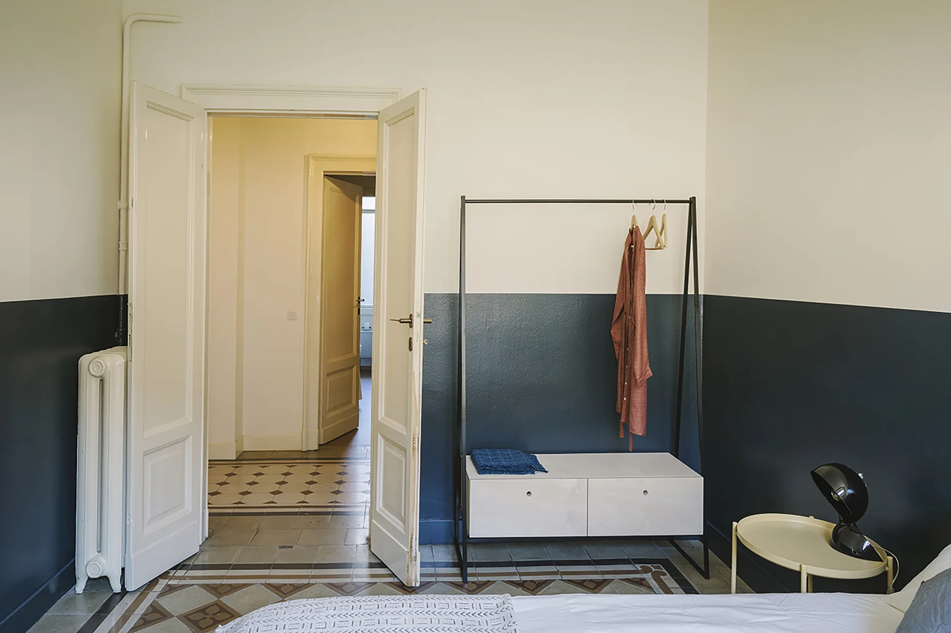 A bedroom featuring white and dark blue painted walls, a black metal clothing rack, patterned tile flooring, and open double doors.