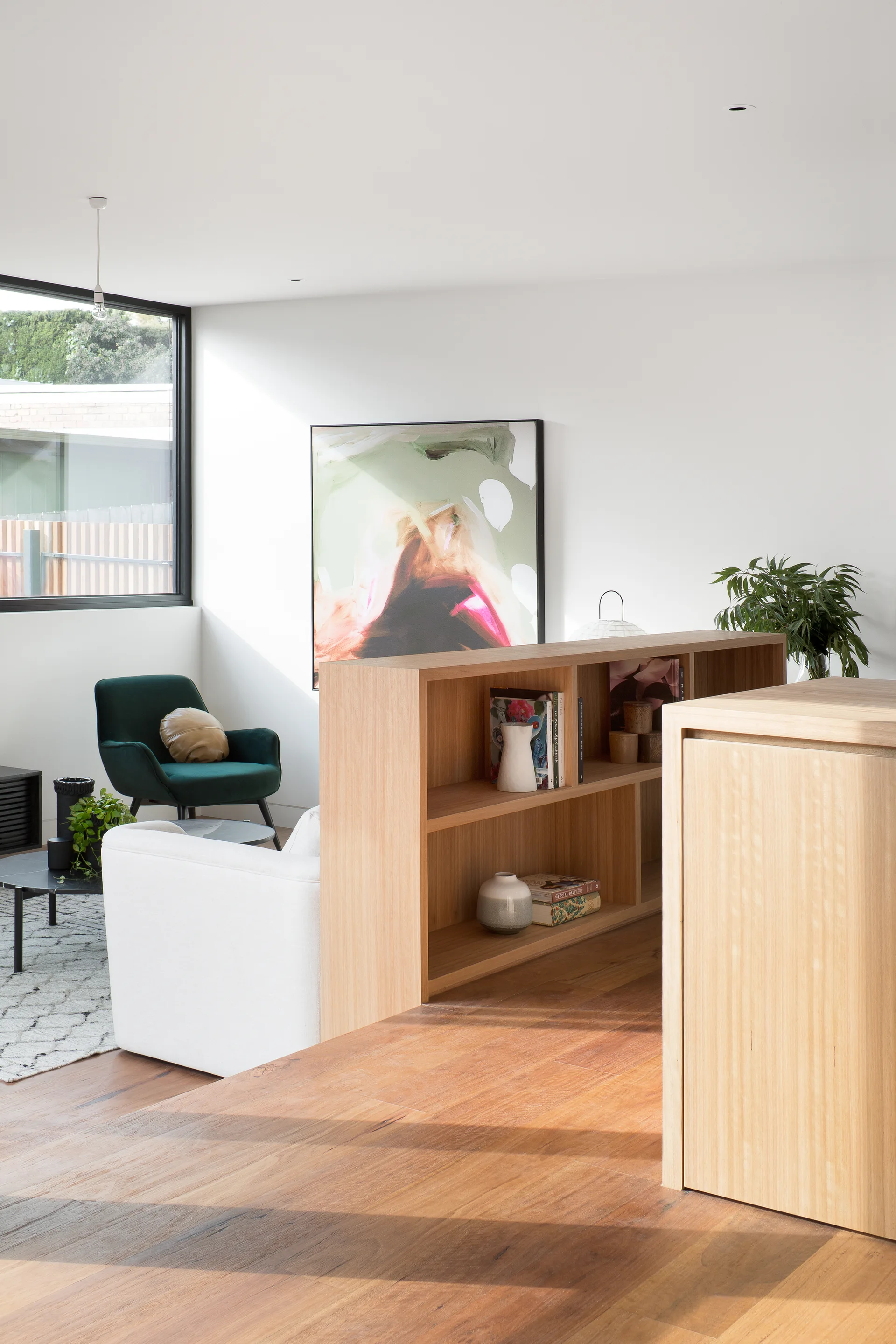 A modern living room features a white sofa, a green velvet armchair, and a low wooden bookshelf serving as a room divider on hardwood floors.