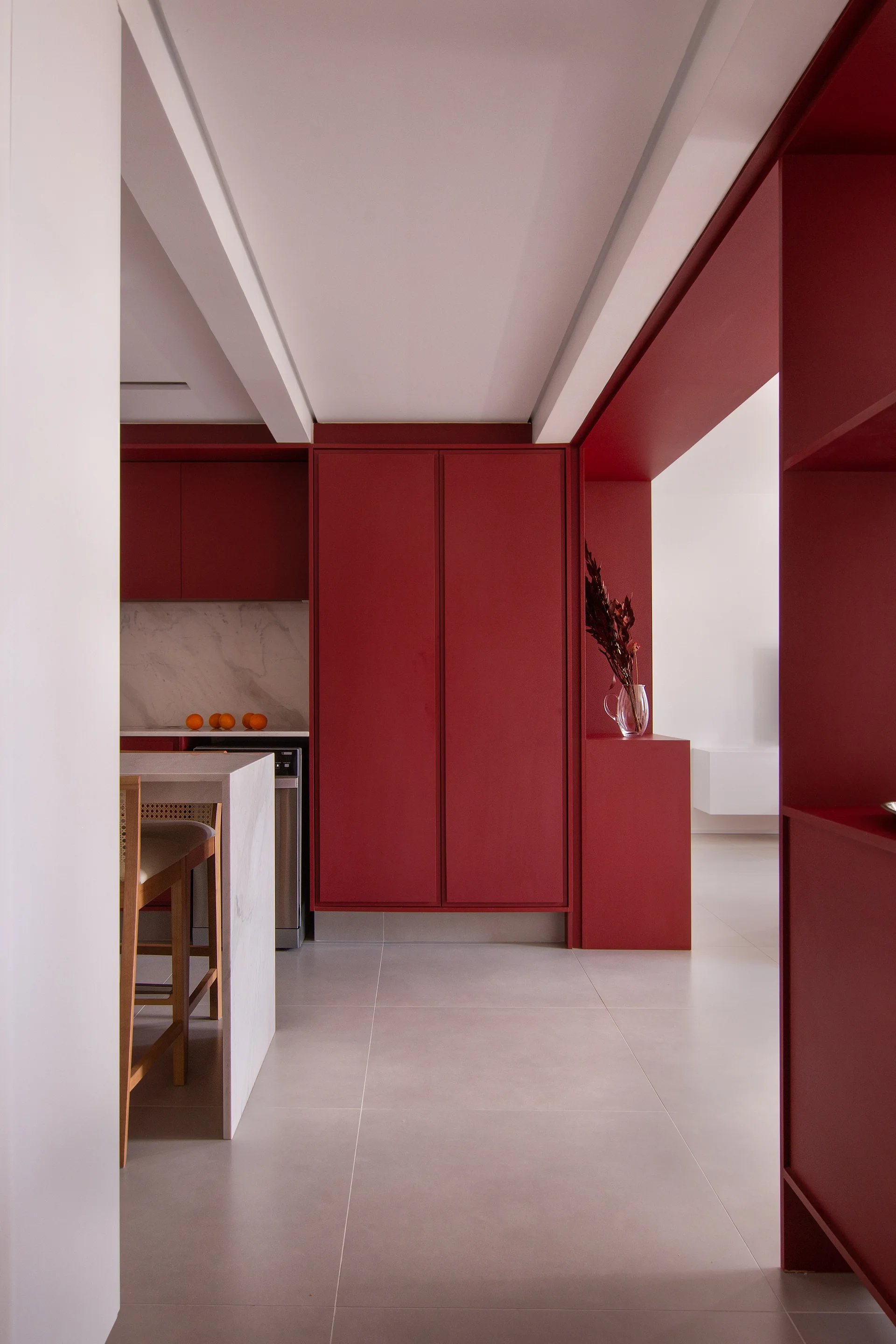 A minimalist kitchen features vibrant matte red cabinetry, a white stone island with cane bar stools, and light grey tile floors.