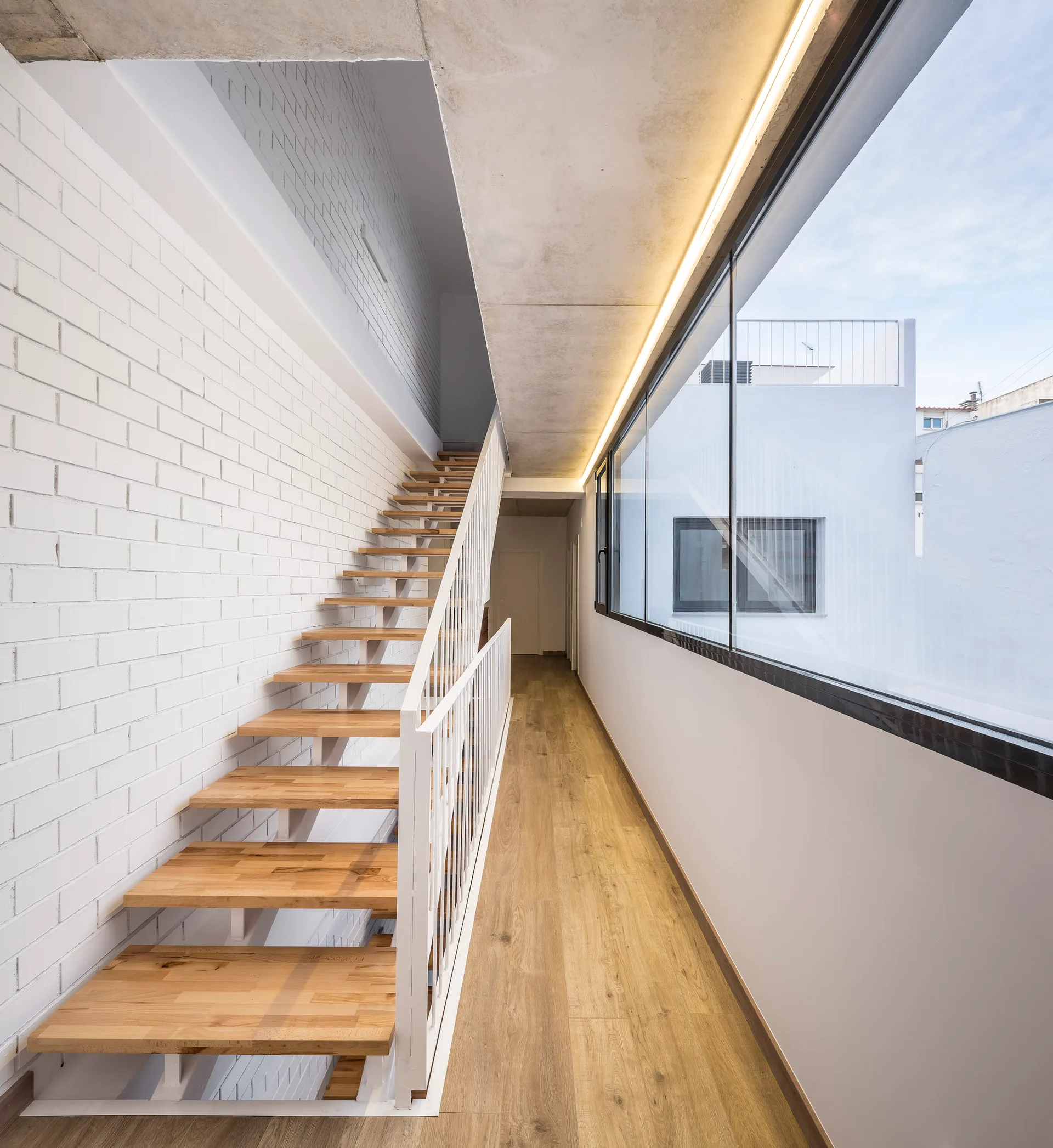 A modern interior corridor features a floating wooden staircase with white railings alongside a painted brick wall and a long horizontal window.
