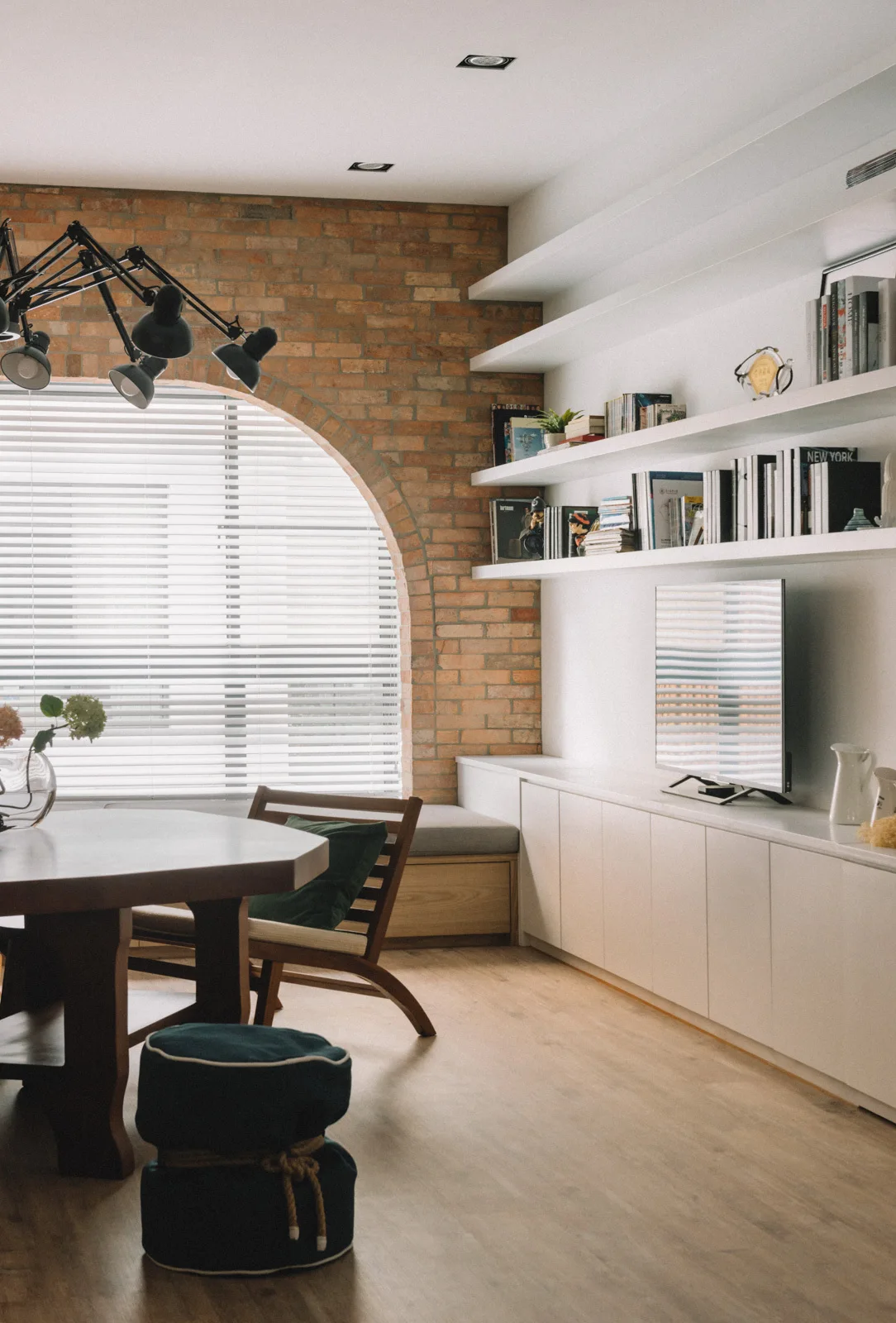 A bright living and dining space features an exposed brick wall with an arched window, white built-in cabinets, and a wooden table.