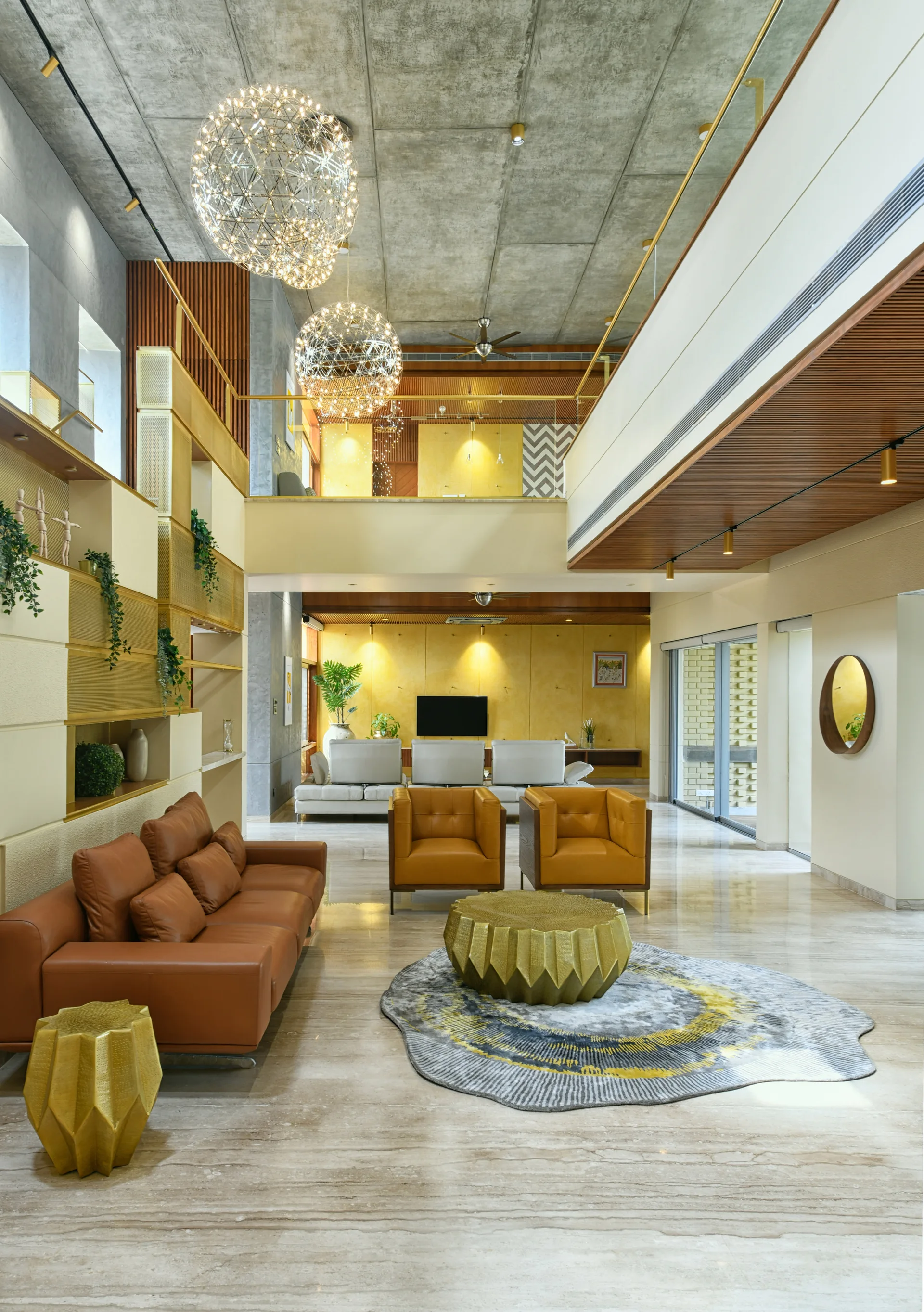 Double-height living room with brown leather seating, faceted gold tables, and spherical wire pendant lights below a concrete ceiling.